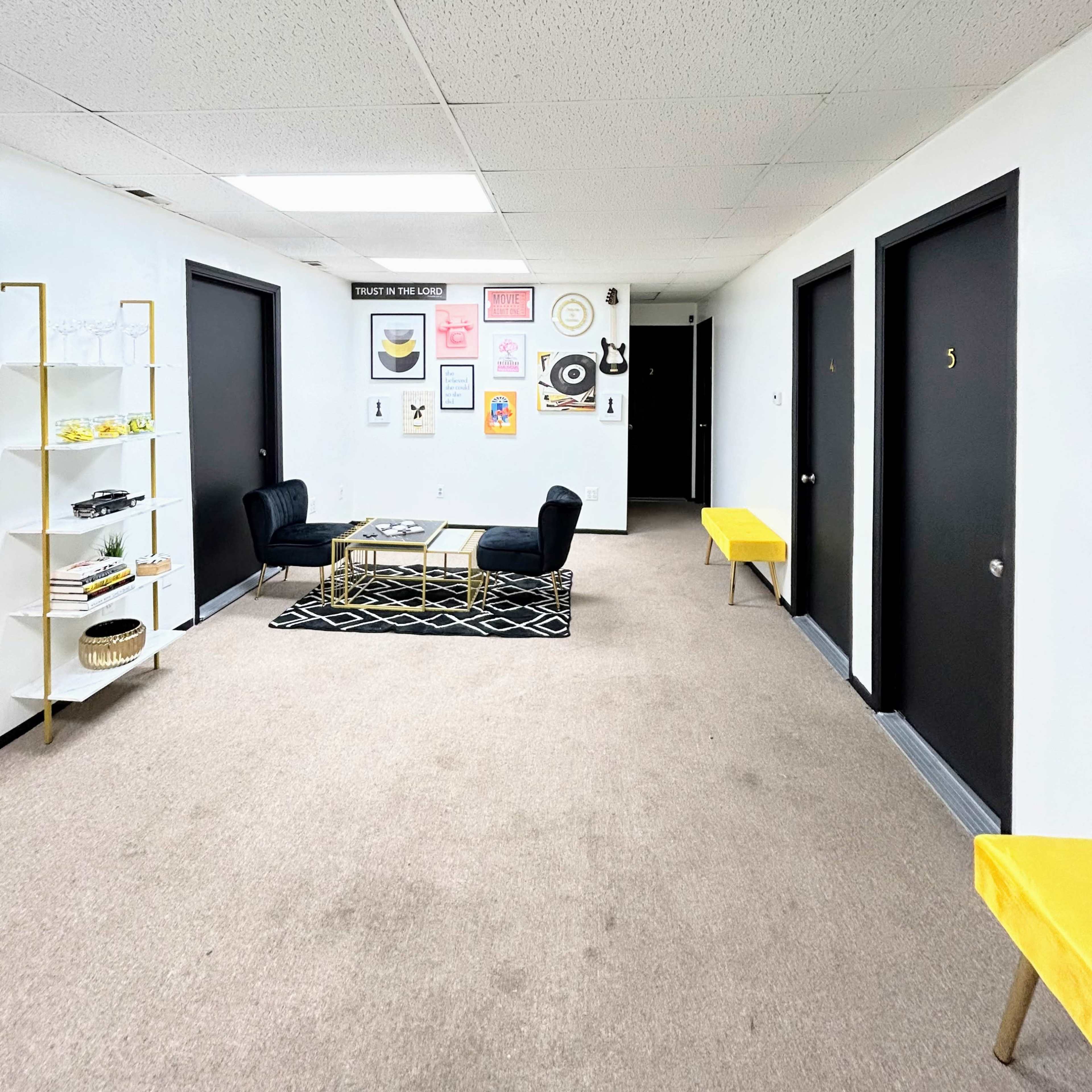 A hallway with four black doors, a coffee table with two black chairs, and art pieces on the wall, all set on a carpeted floor.