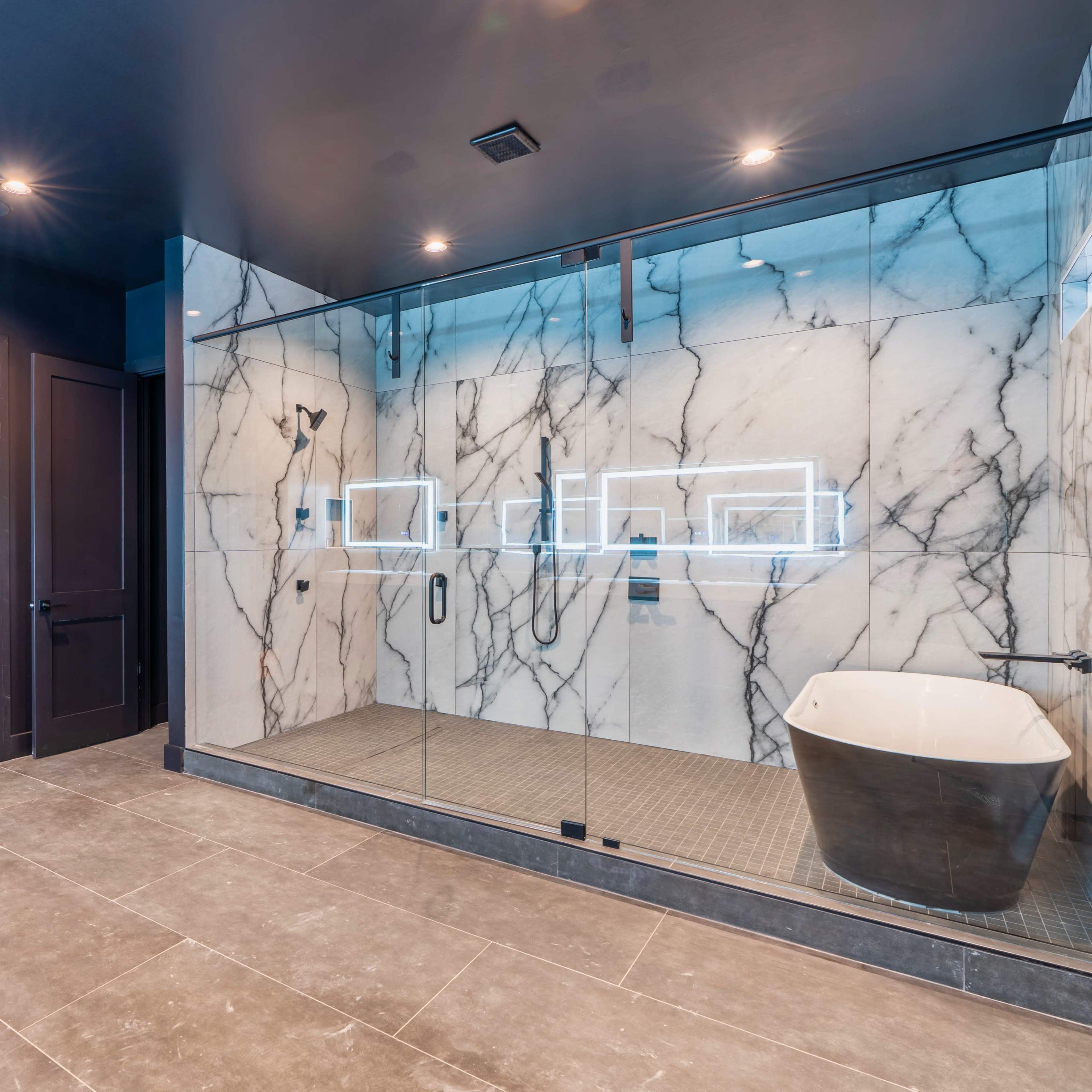 A modern bathroom featuring a glass-encased shower with marble walls, a freestanding black tub, and dark cabinetry.