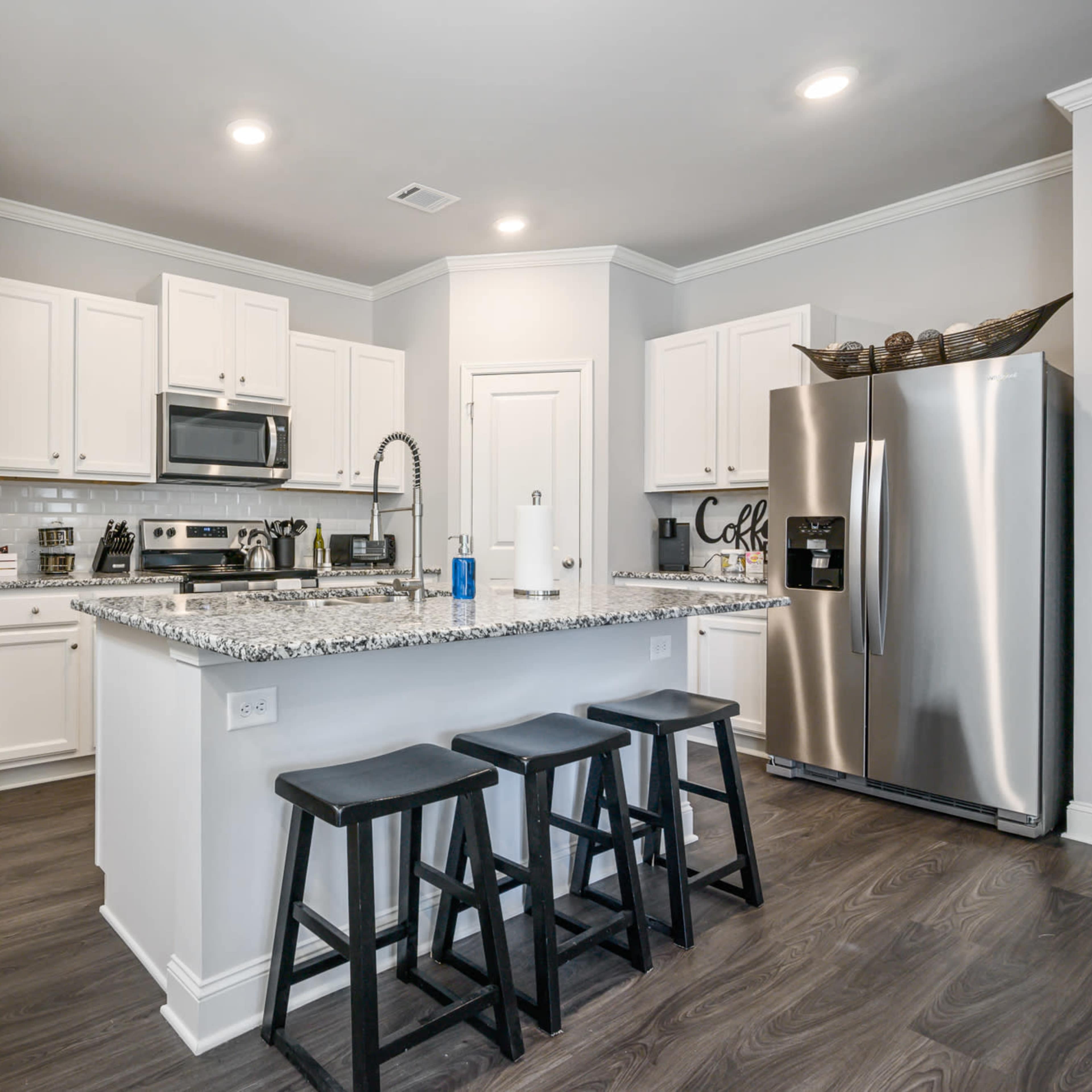 A modern kitchen with white cabinets, stainless steel appliances, and a granite countertop with four black stools at the island.