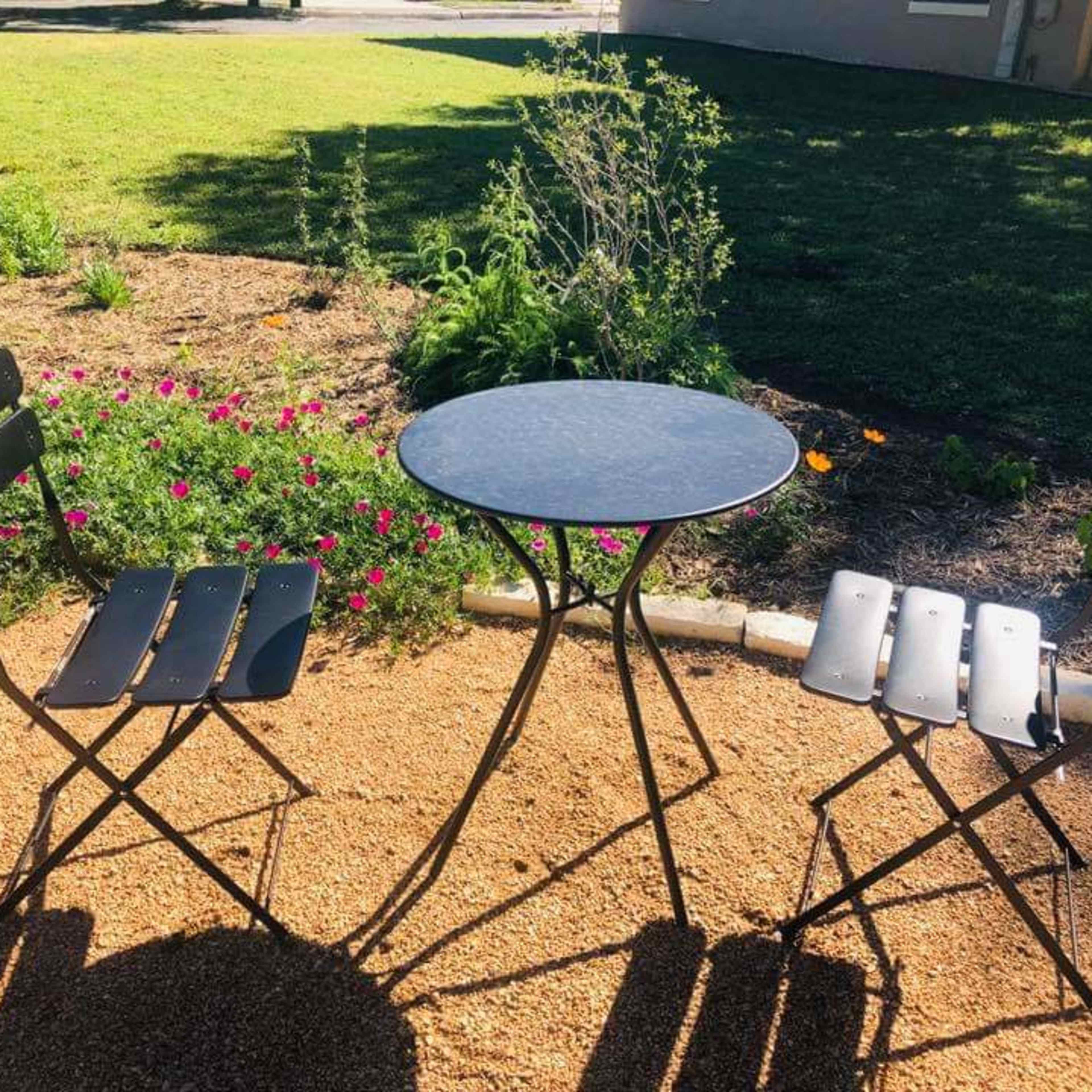 A small round table is positioned between two folding chairs on a gravel patch surrounded by flowers and greenery.
