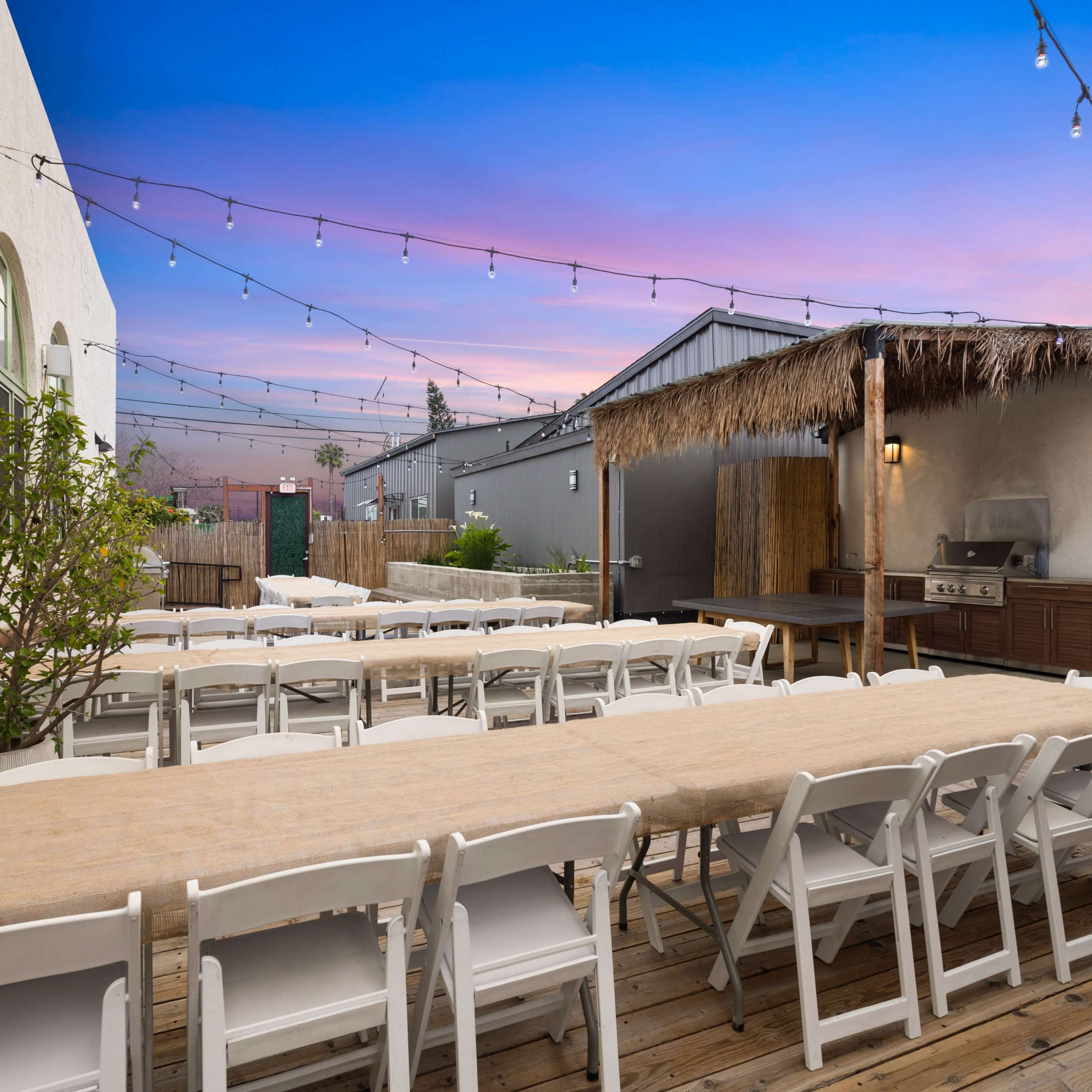 A well-arranged outdoor seating area with long tables set up under string lights, adjacent to a barbecue area and surrounded by a landscaped space at dusk.
