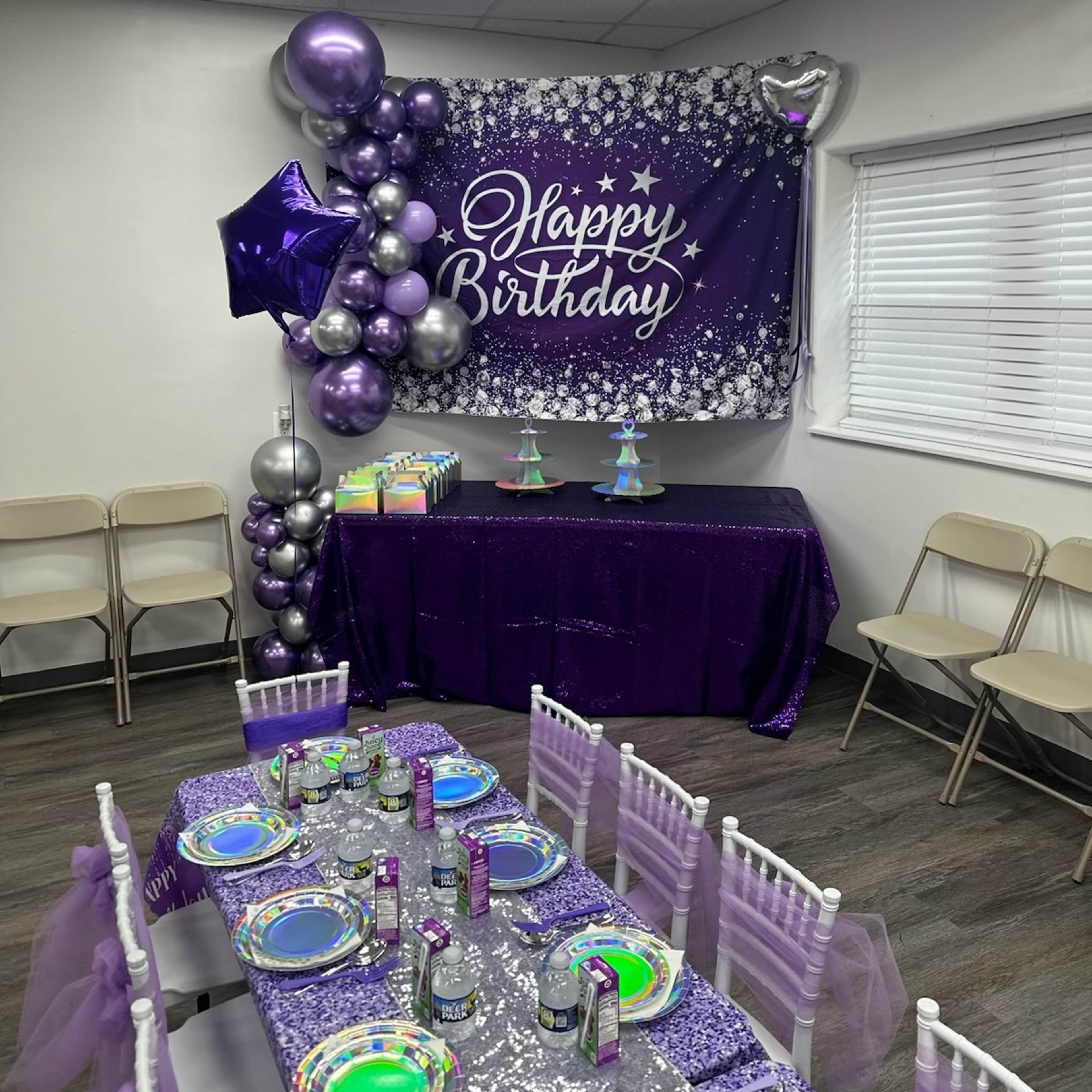 A birthday celebration setup featuring a "Happy Birthday" banner, purple decorations, a table with colorful plates, and a dessert display.