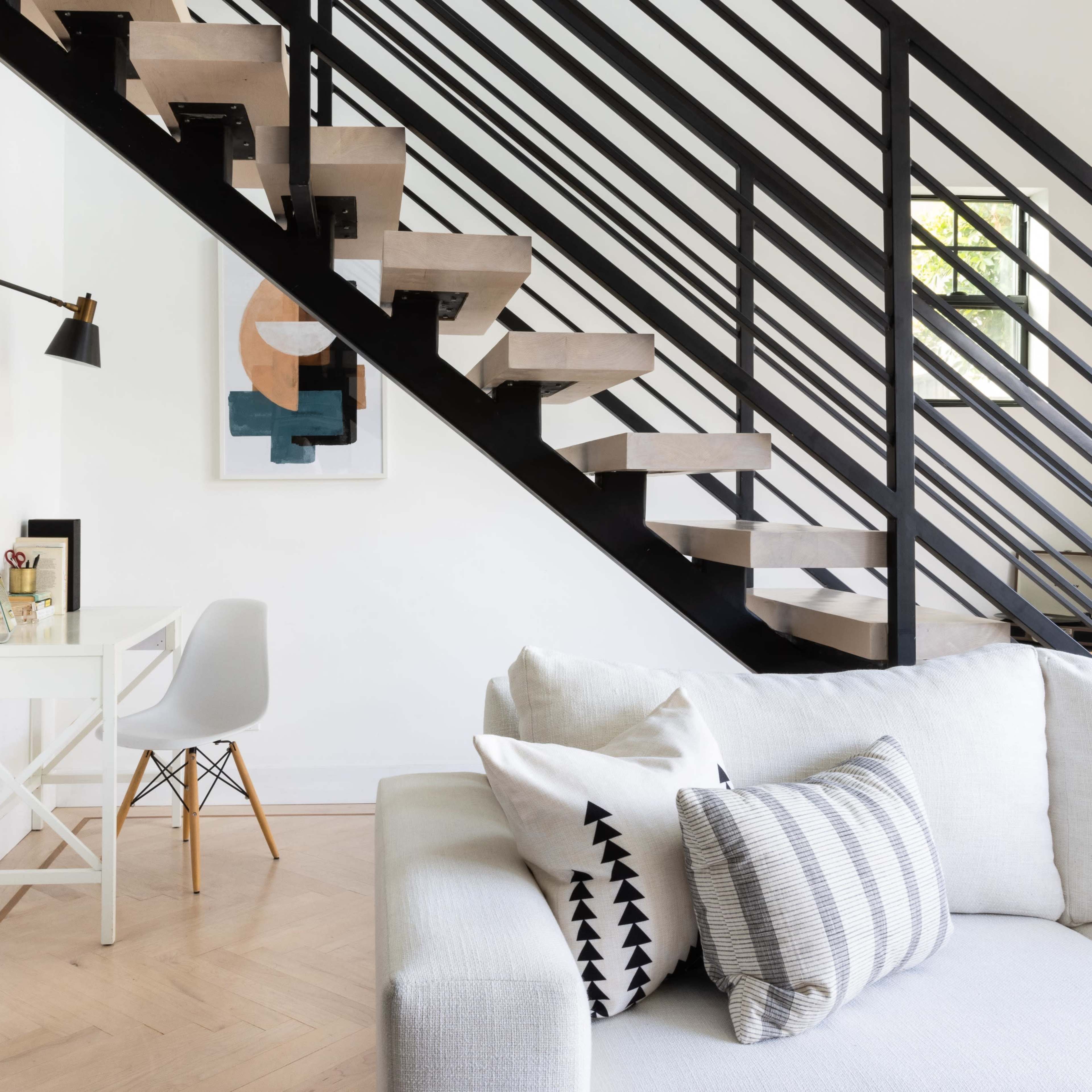 A modern living room features a white sofa, a minimalist desk under a staircase, and a framed piece of artwork on the wall.