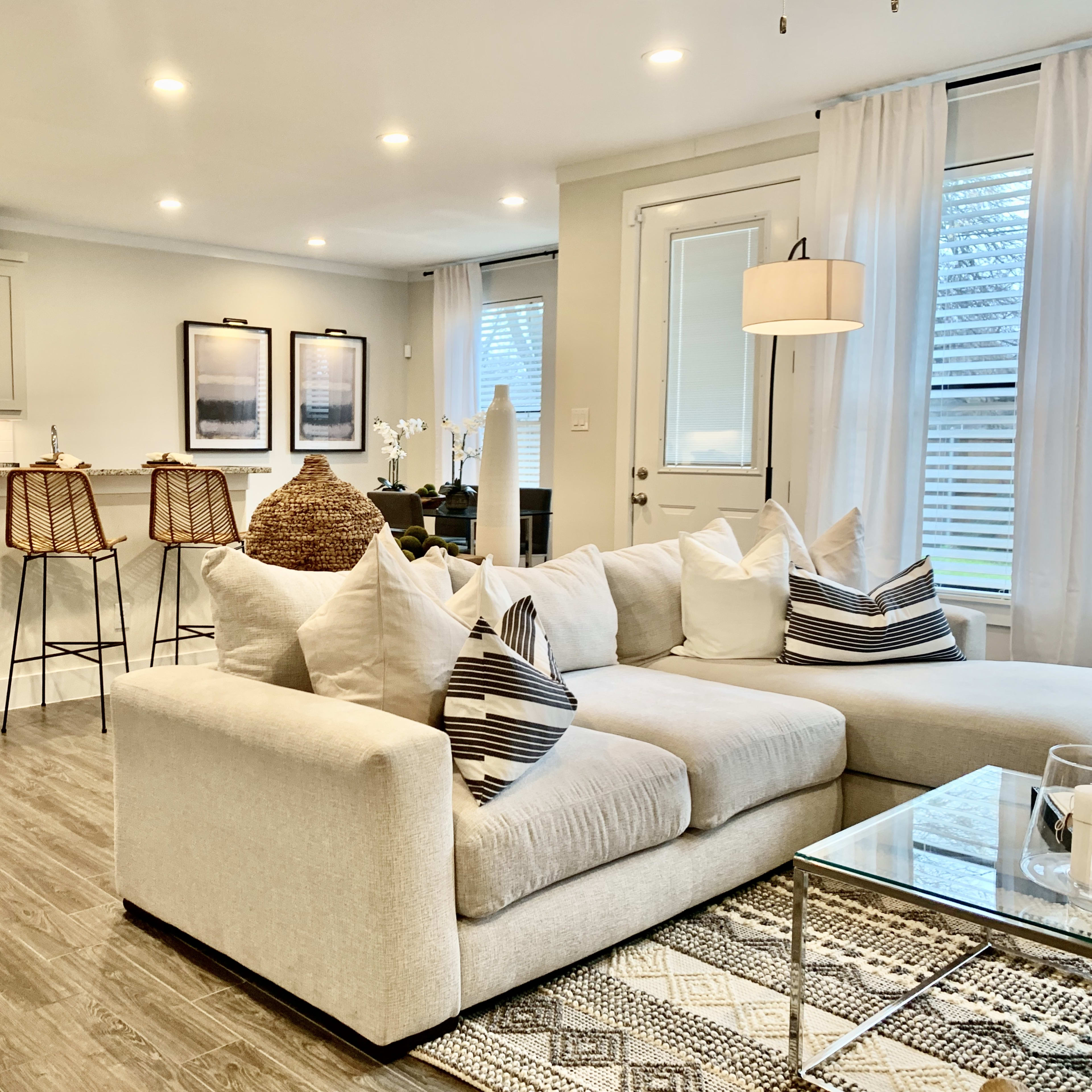 A modern living space with a light-colored sectional sofa, a kitchen area featuring bar stools, and large windows with sheer white curtains.