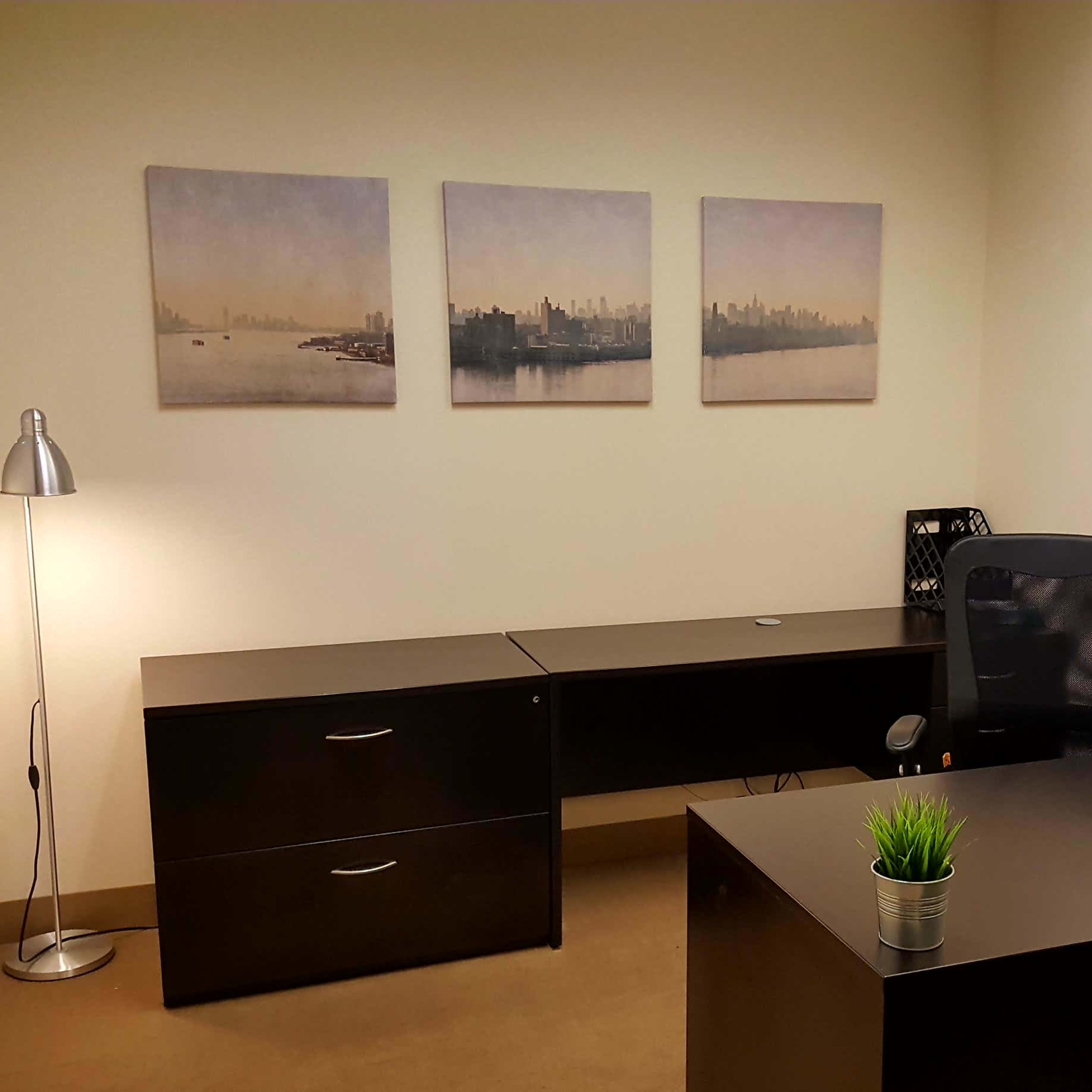 A minimalistic office space featuring two black desks, a chair, a lamp, a telephone, and three framed cityscape prints on the wall.