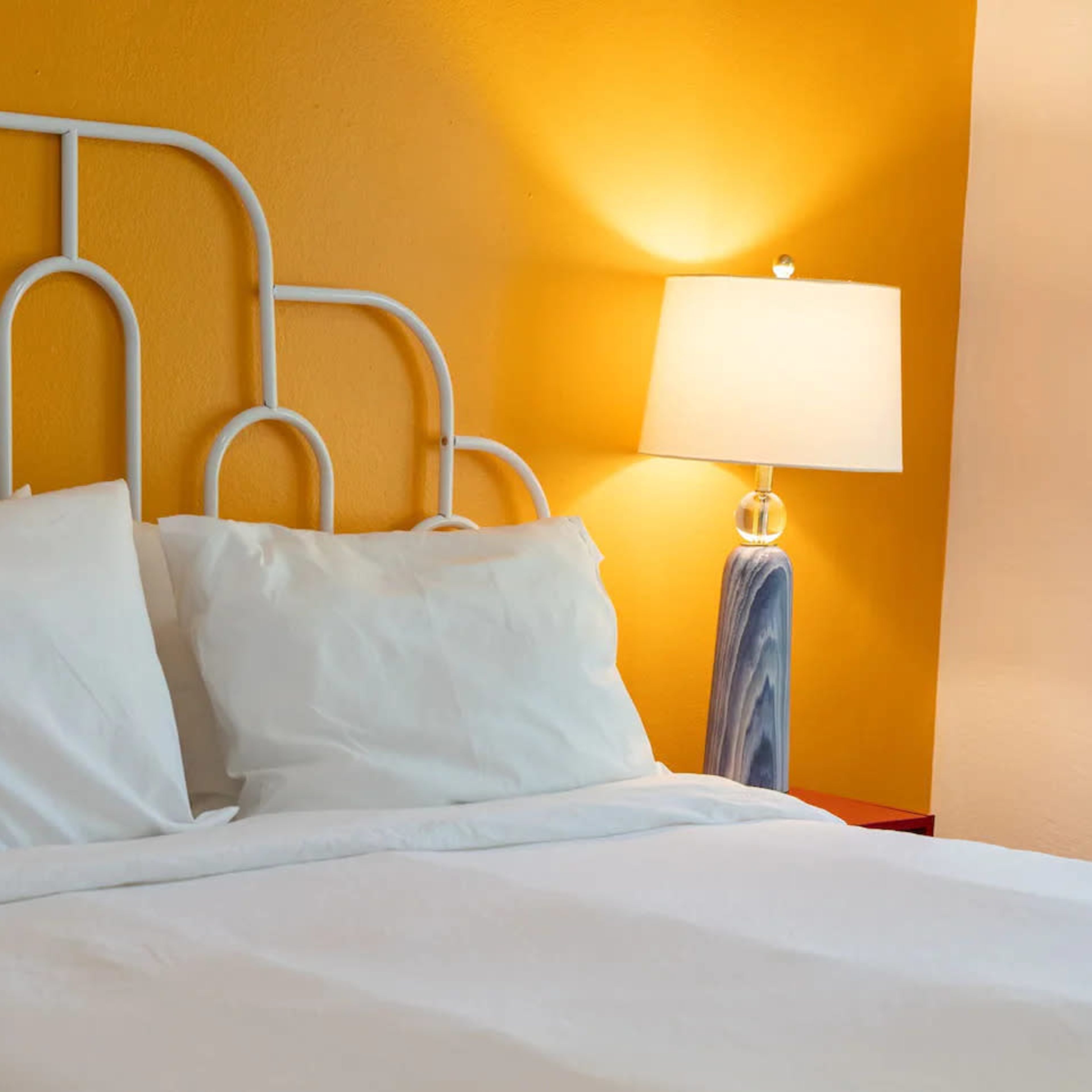 A neatly made bed with white bedding against a bright yellow wall, accompanied by a table lamp with a marbled base.