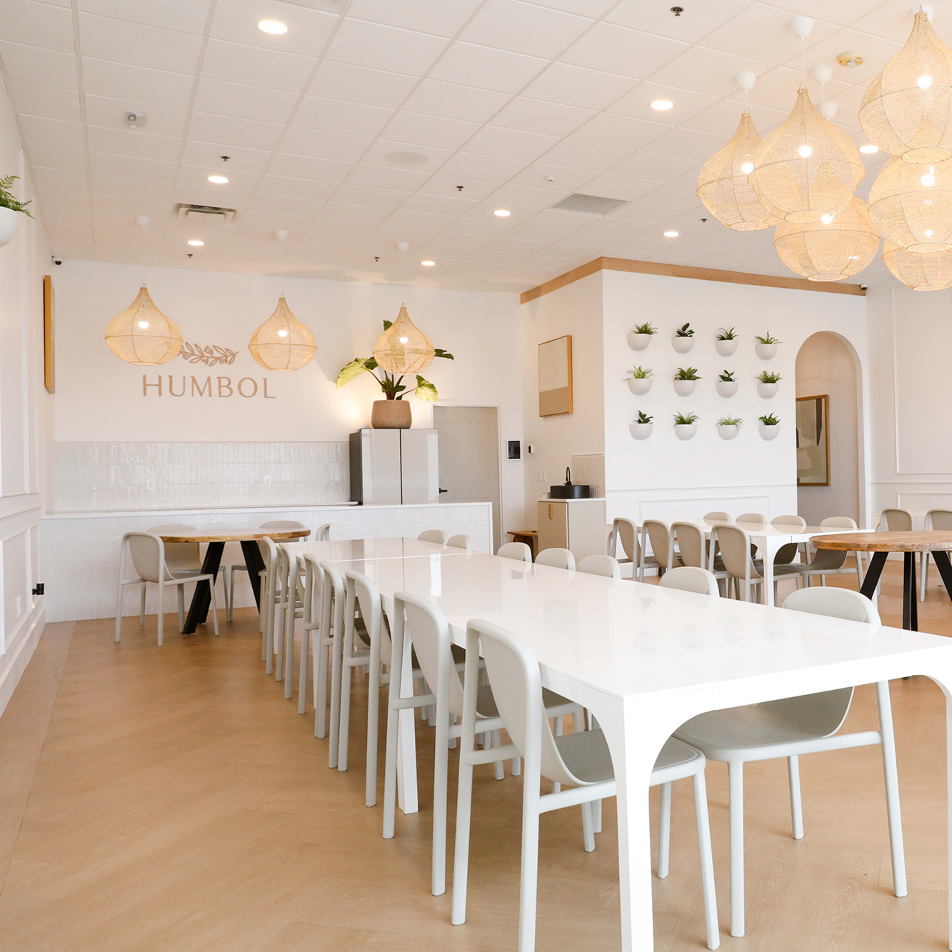 A modern, well-lit dining area with white tables and chairs, featuring decorative pendant lights and greenery on the walls.
