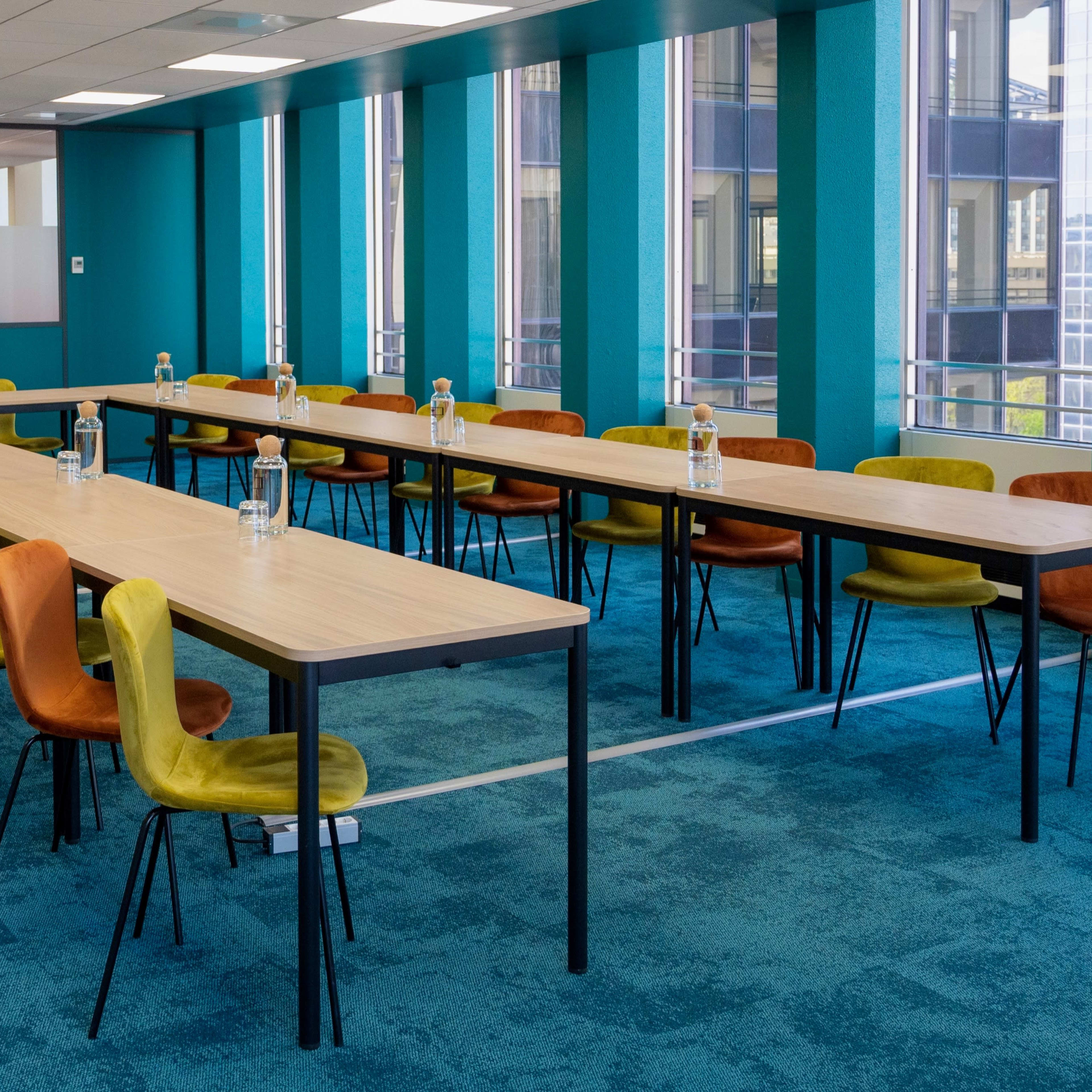 A spacious meeting room features long tables arranged in a row, each accompanied by colorful chairs and bottles of water.