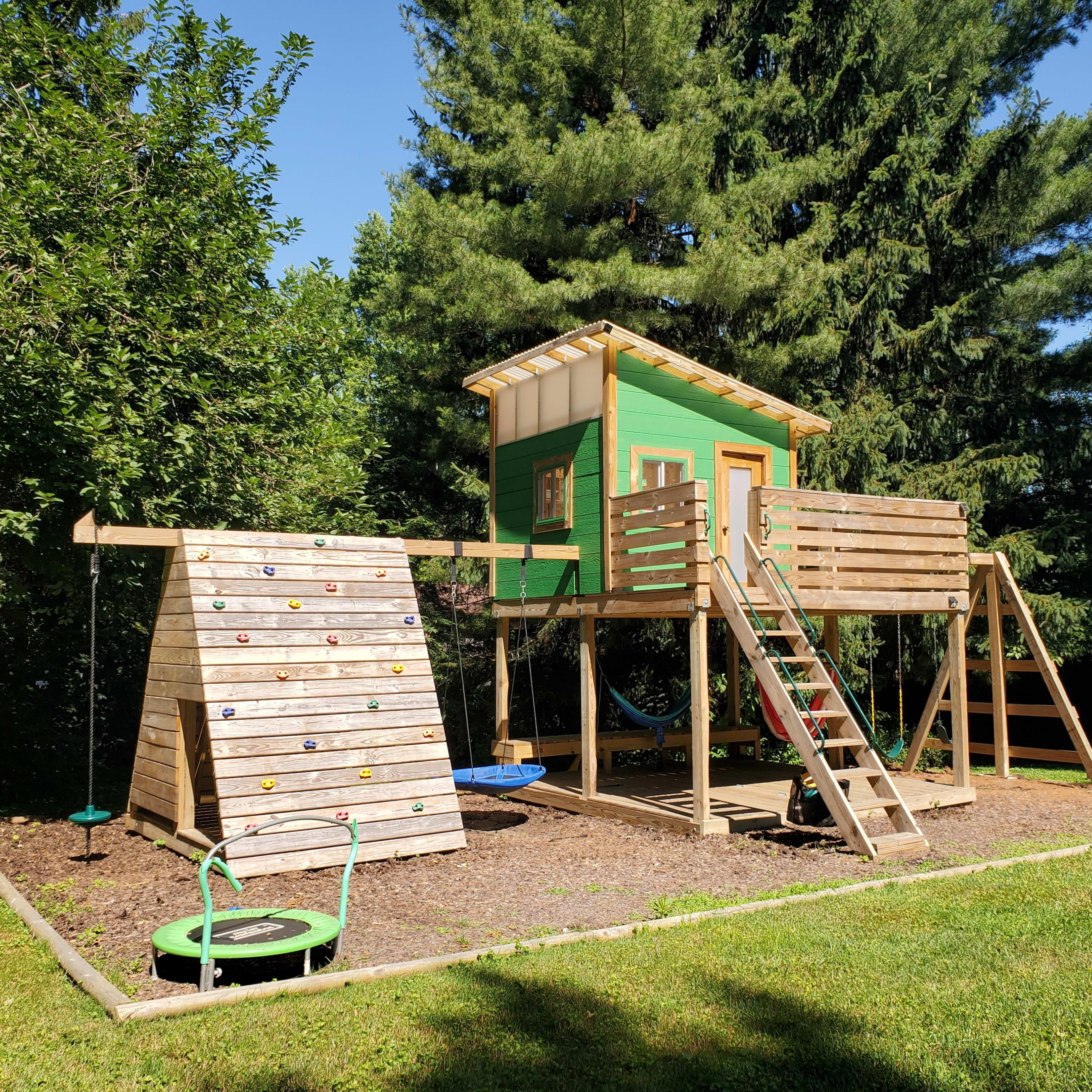A wooden playset with a green treehouse, a climbing wall, and a swing set, all situated in a grassy yard surrounded by trees.