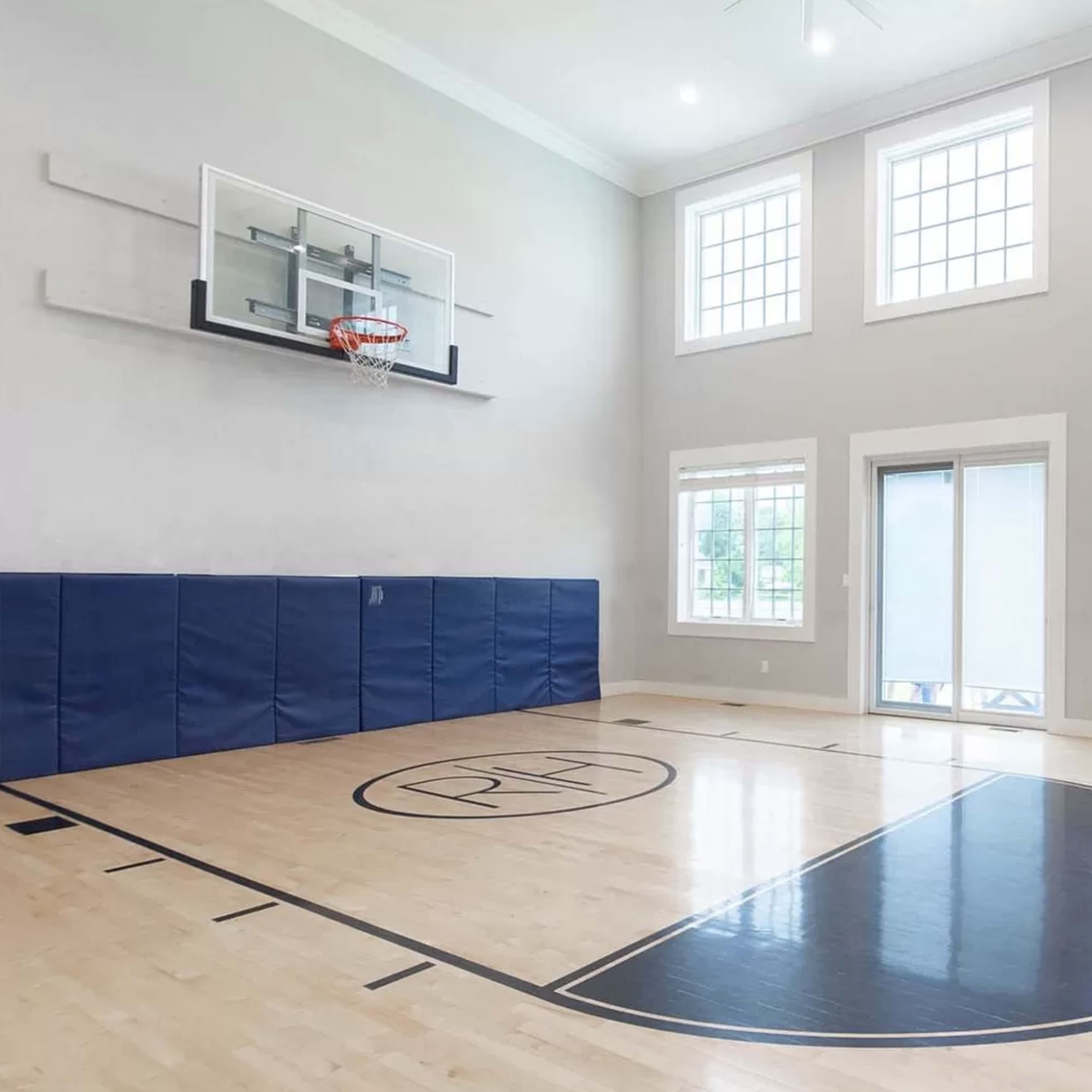 An indoor basketball court with a hoop, large windows, and light wood flooring.