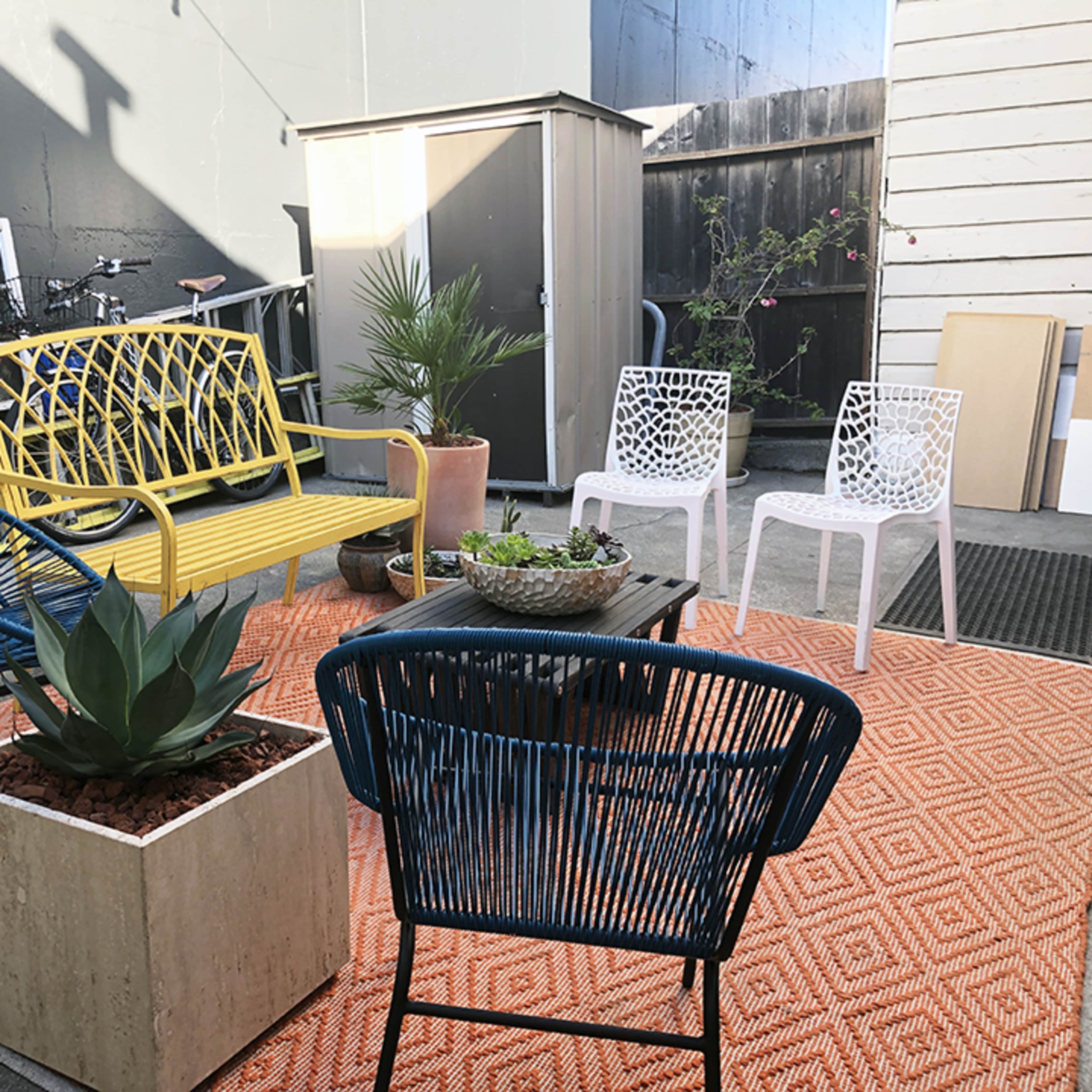 A small outdoor seating area featuring a yellow bench, three white chairs, and two blue armchairs arranged around a central table on a patterned rug.