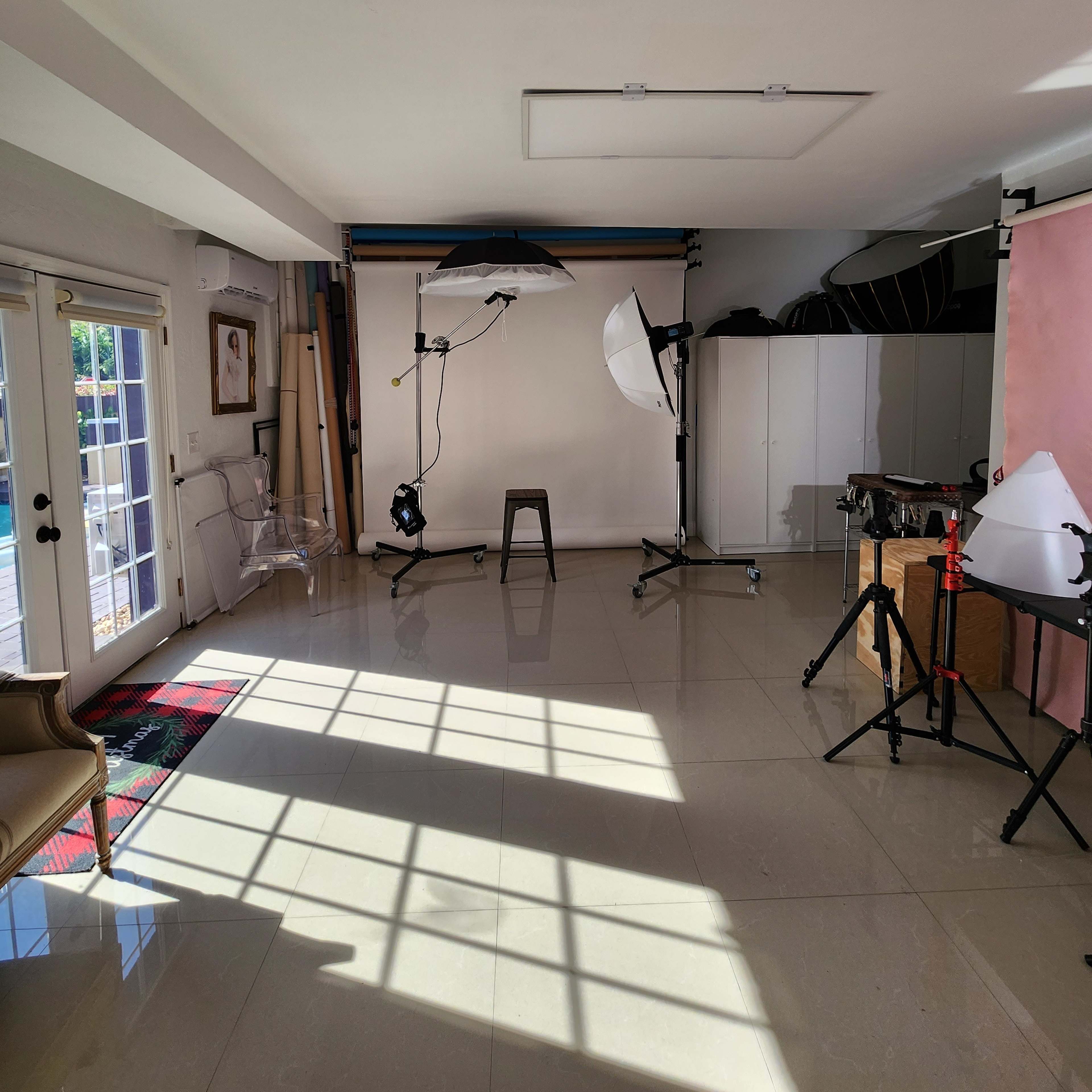 A photography studio features a blank backdrop with lighting equipment and a wooden stool on glossy tile flooring.
