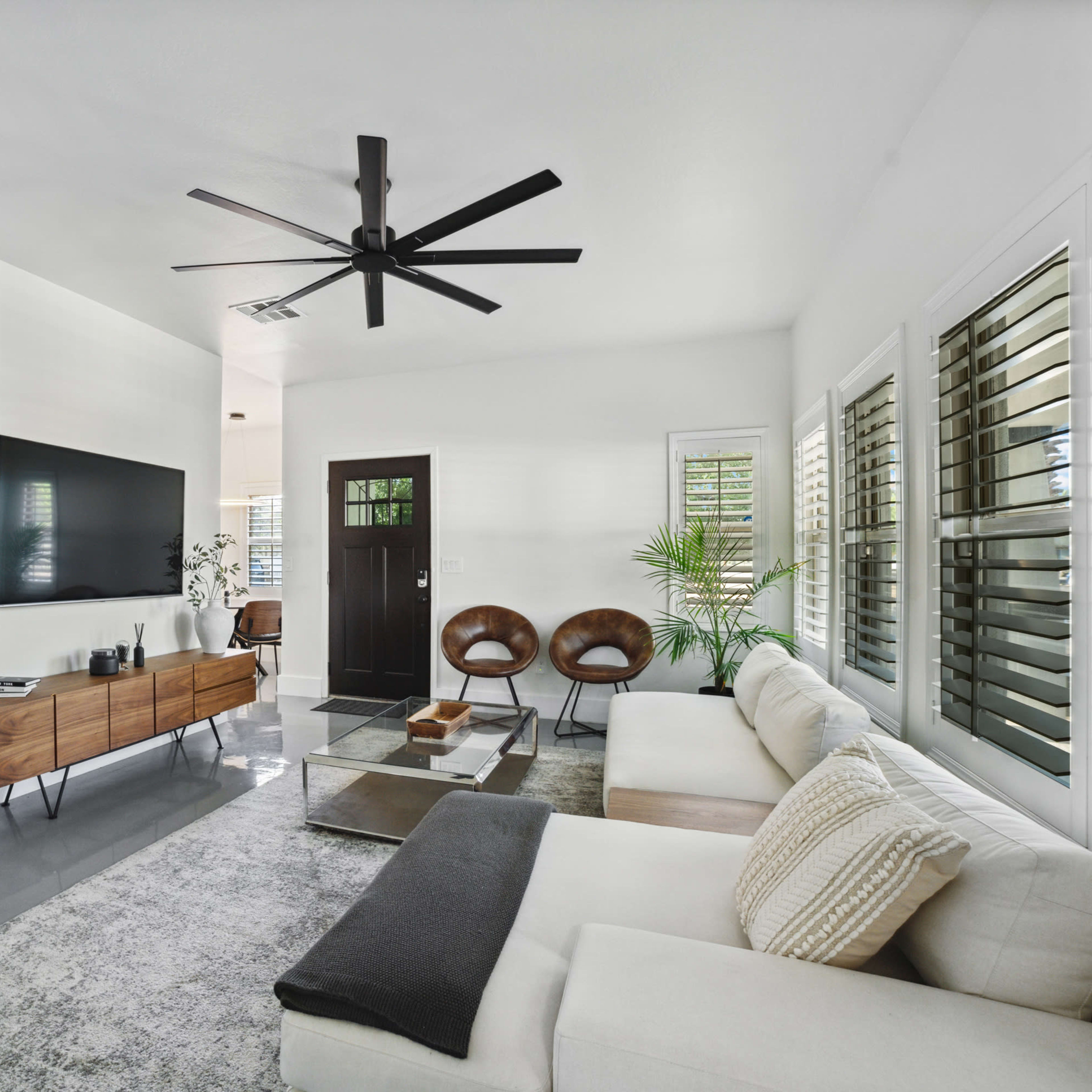 The living room features a white color scheme with a large television mounted on the wall, a sleek sofa, two brown chairs, and a potted plant near a black front door.