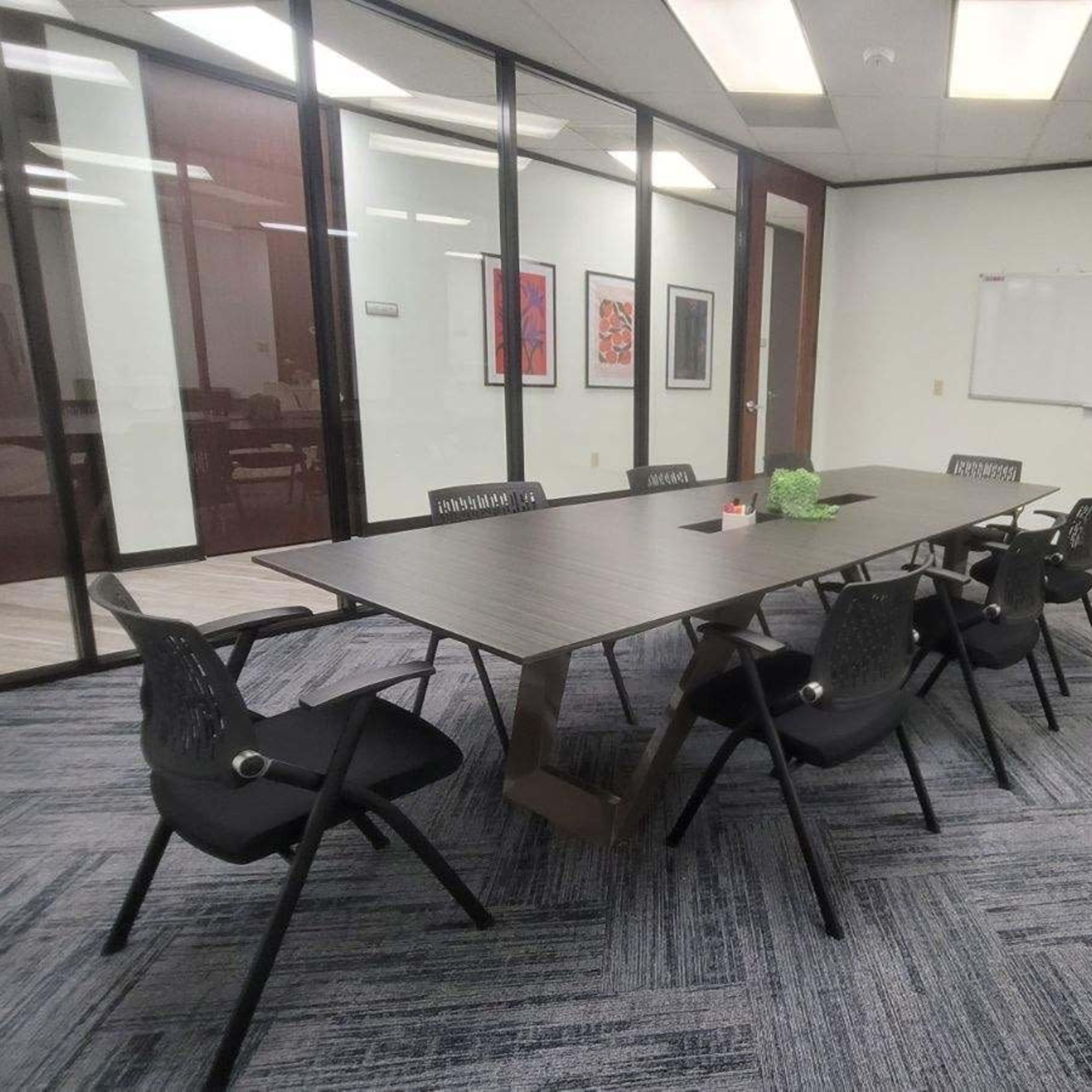 A modern conference room features a long table surrounded by black chairs, with glass walls and a whiteboard in the background.
