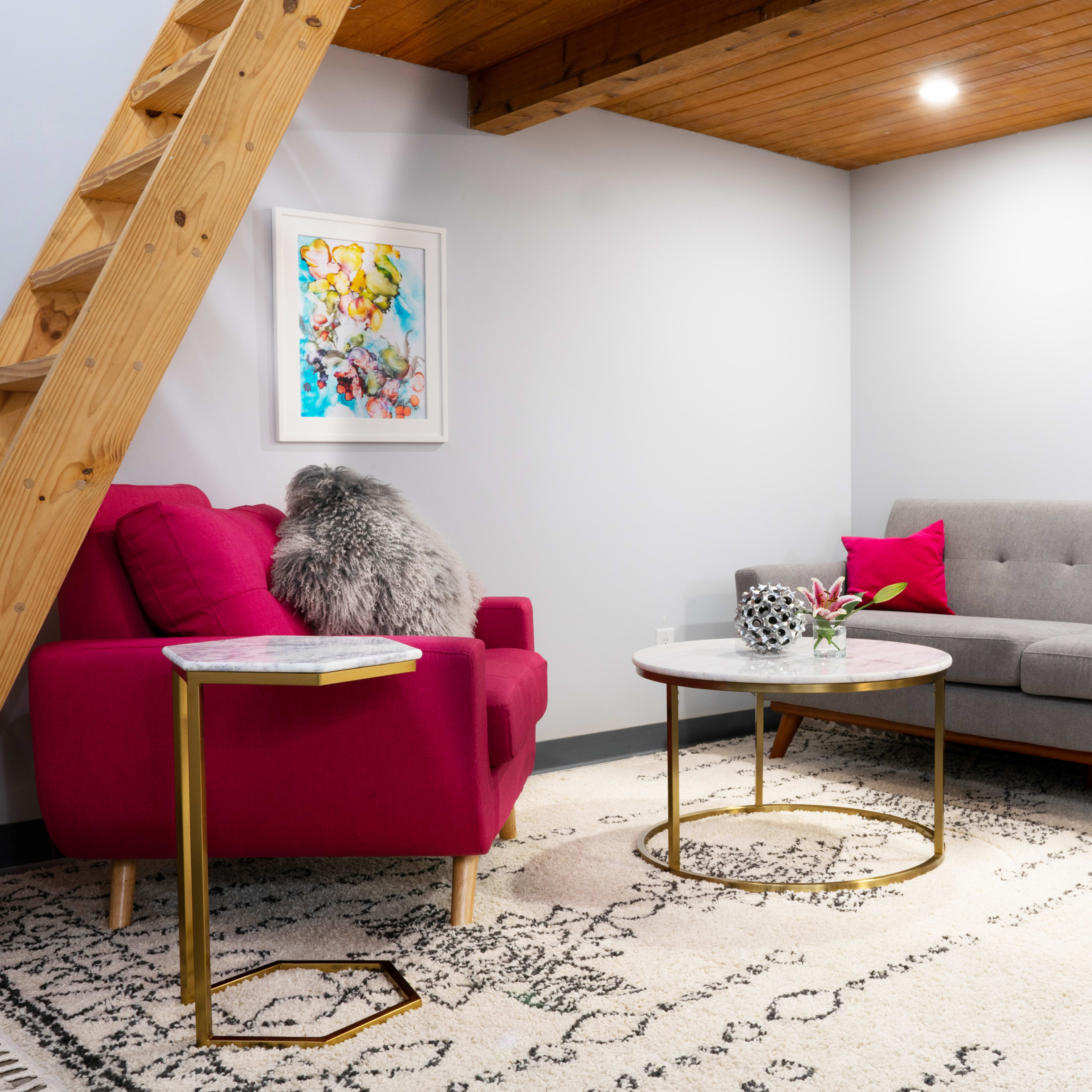 A modern living area featuring a gray couch, a pink chair, a marble coffee table, and a patterned rug under a wooden staircase.
