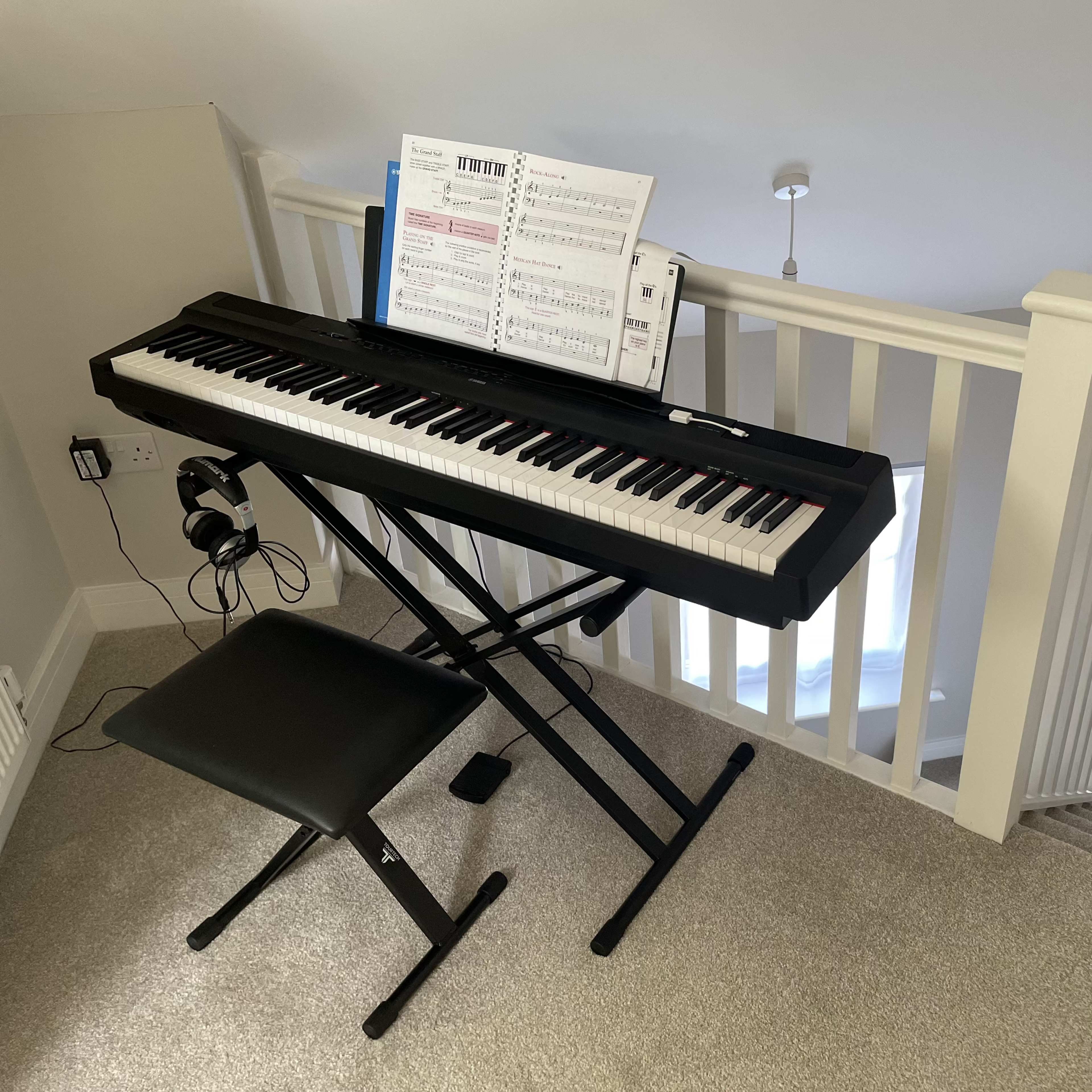 A digital piano with sheet music and headphones is positioned on a carpeted stair landing near a white railing overlooking a staircase.