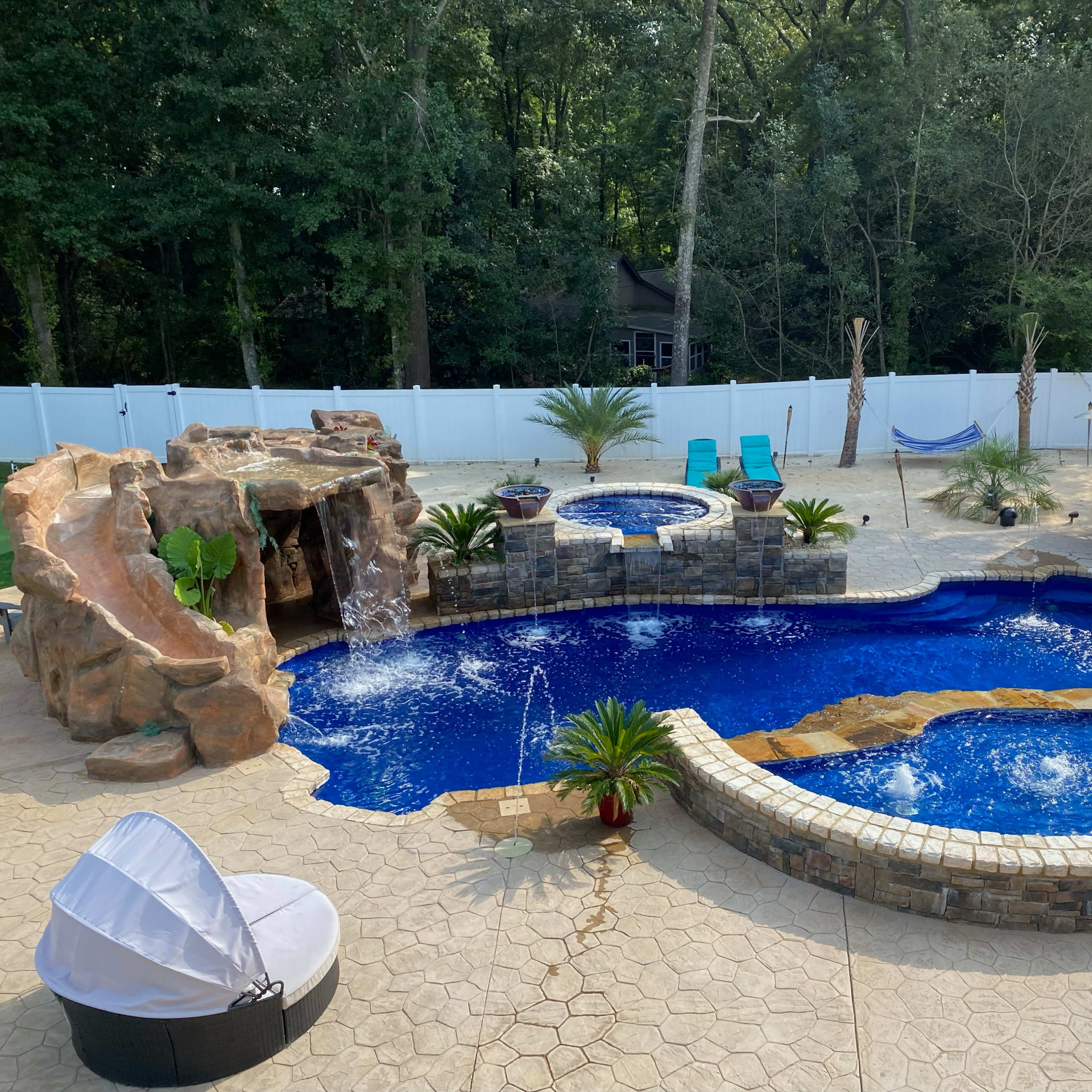A backyard pool area featuring a large, curved swimming pool with a rock waterfall, a hot tub, lounge chairs, and landscaped greenery.