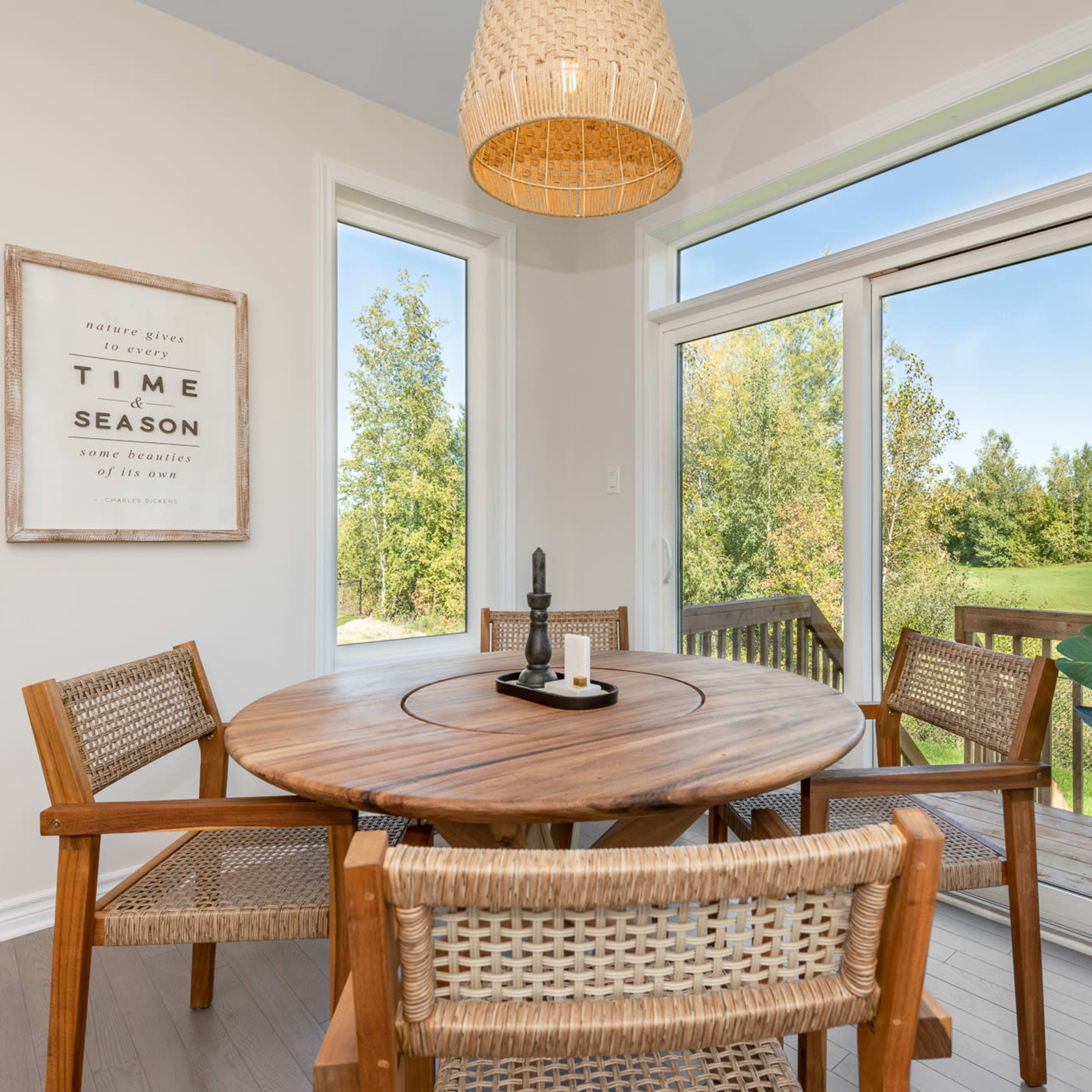 A round wooden dining table with four chairs is set near large windows overlooking a green landscape.