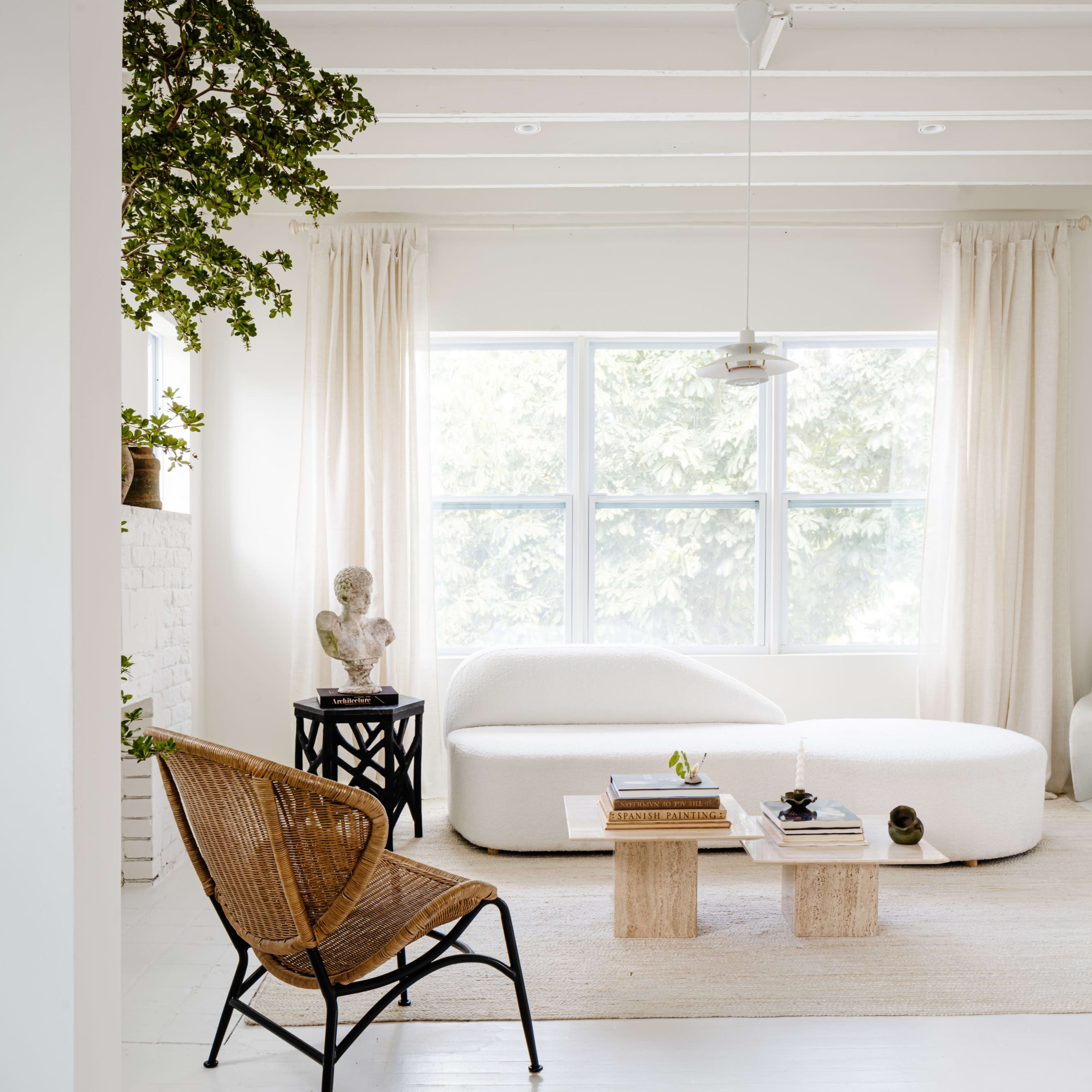 A bright, minimalist living room features a white sofa, a wooden coffee table, a rattan chair, and large windows with sheer curtains.