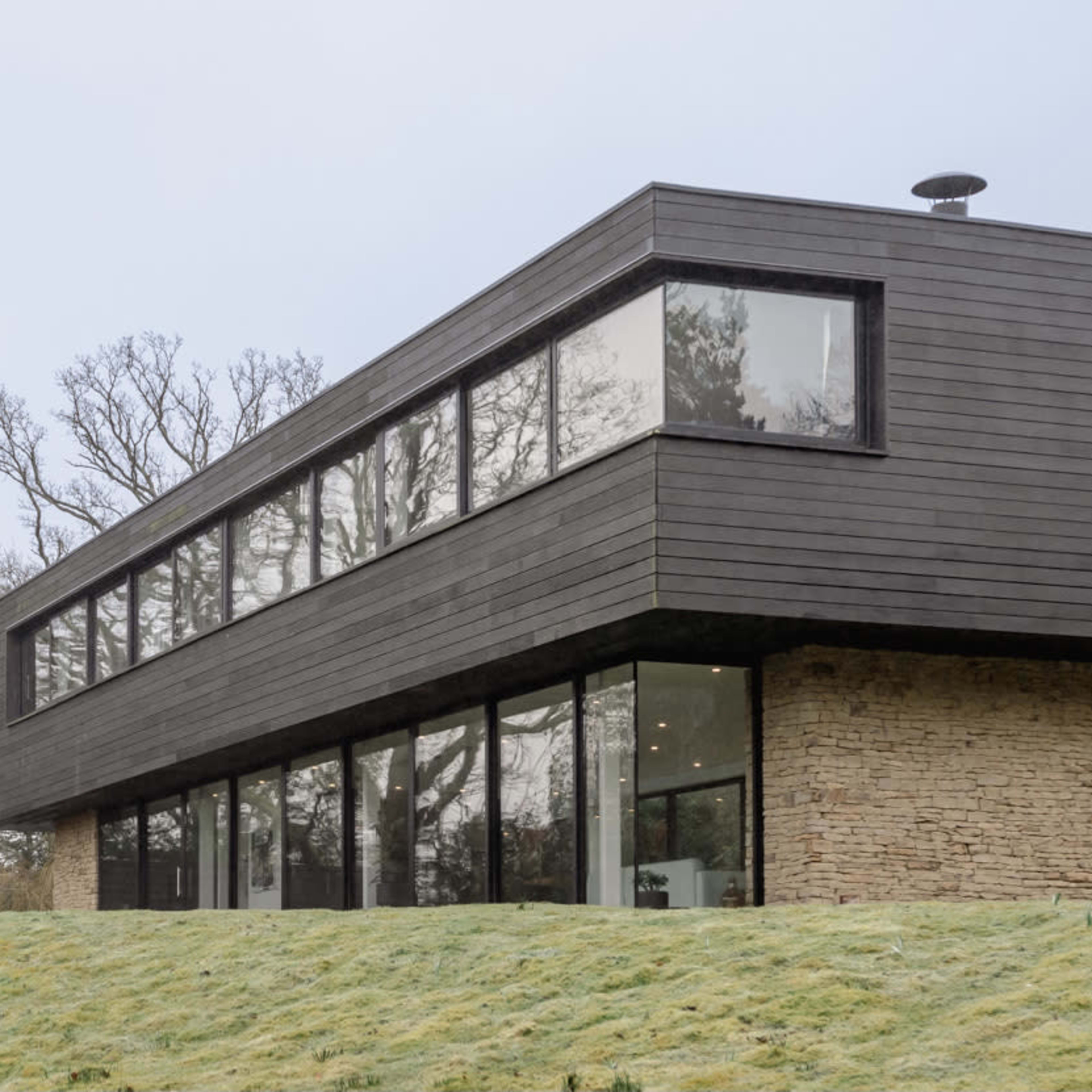 A modern two-story house with a combination of stone and black wood siding is situated on a gently sloped grassy area.