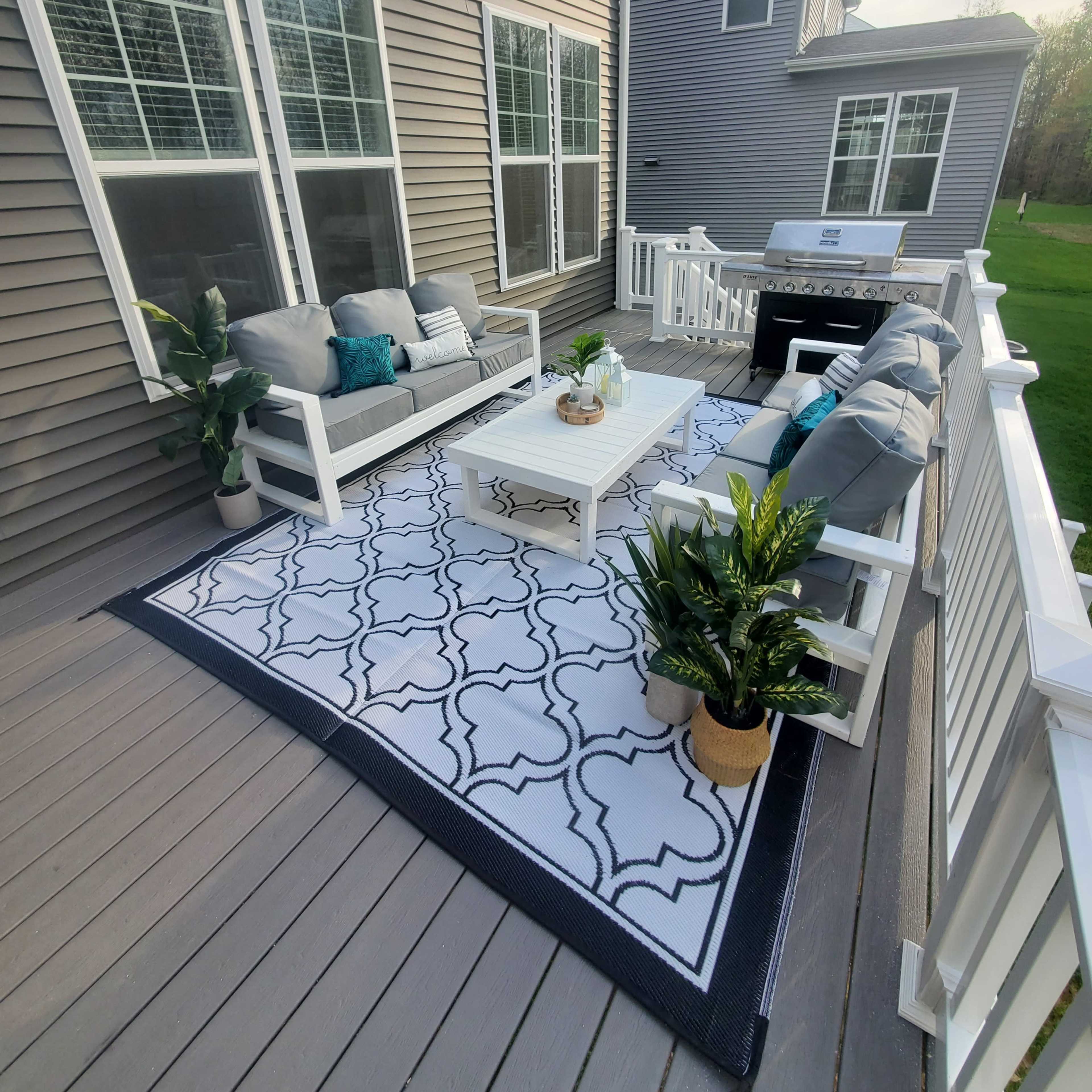 A patio with a seating area featuring a white table, gray couches, decorative pillows, and potted plants on a patterned rug.