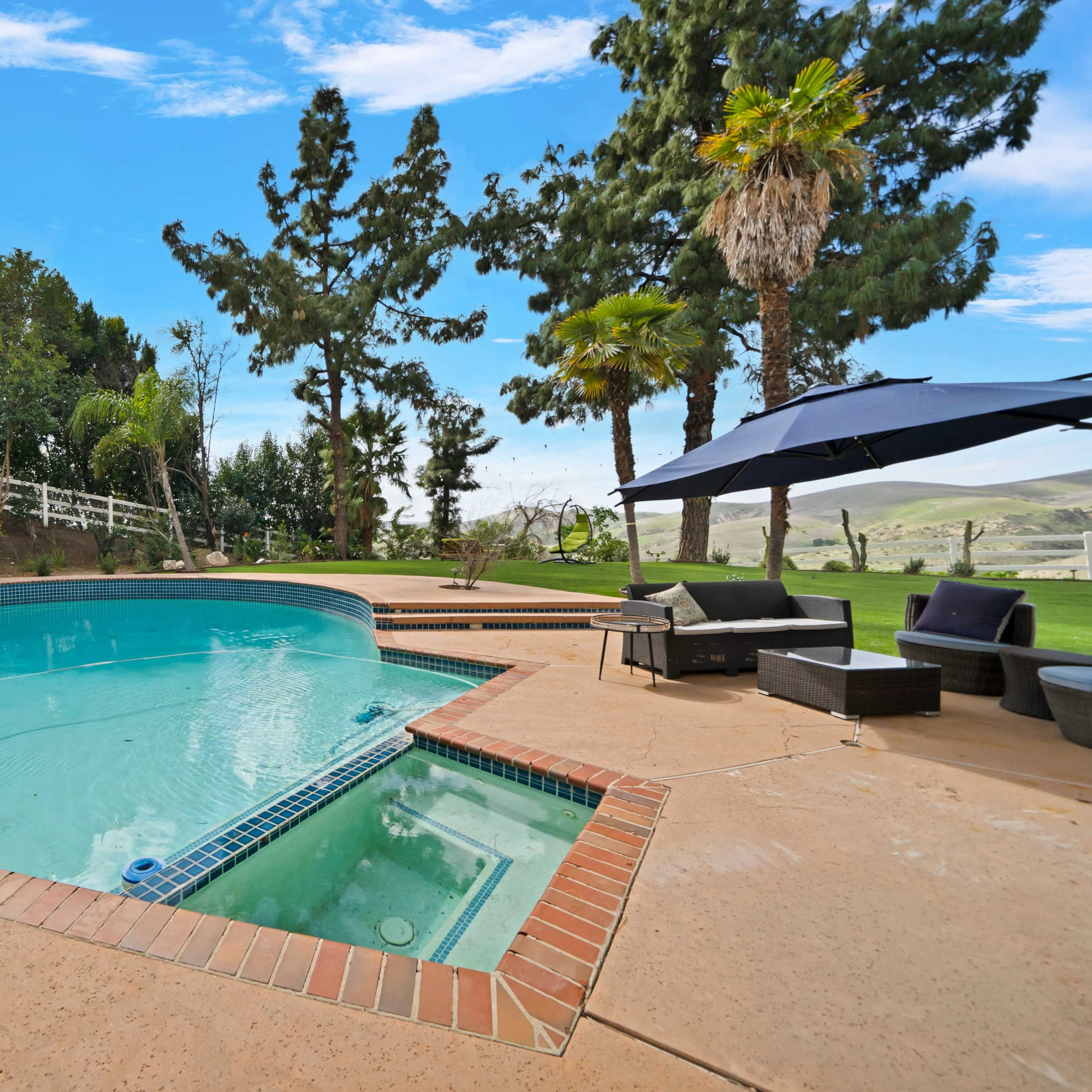 A landscaped backyard featuring a swimming pool with a hot tub, surrounded by palm trees and outdoor furniture under an umbrella.