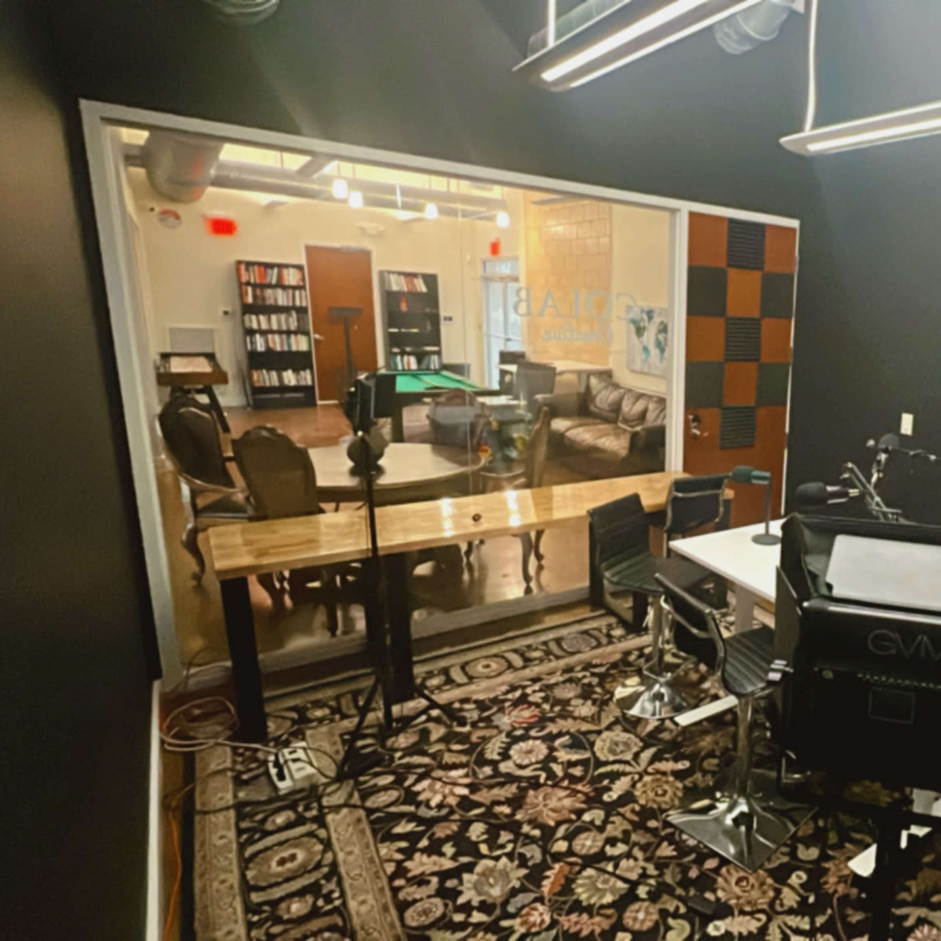 A modern recording studio with a large desk, chairs, and cameras facing a glass-walled lounge area featuring a pool table and bookshelves.