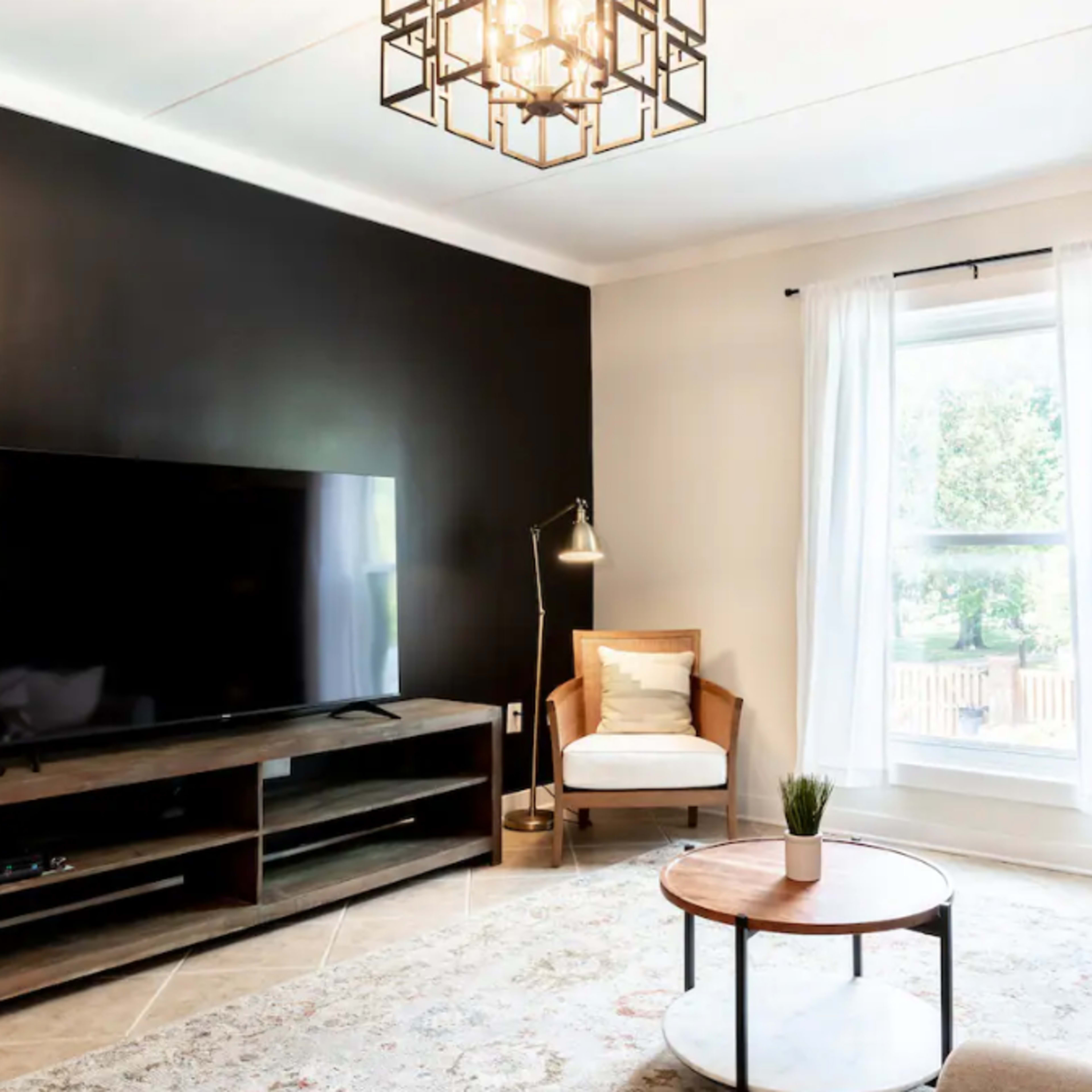 The living room features a television on a wooden stand, a light-colored sofa, a round coffee table, and a large window with sheer curtains.