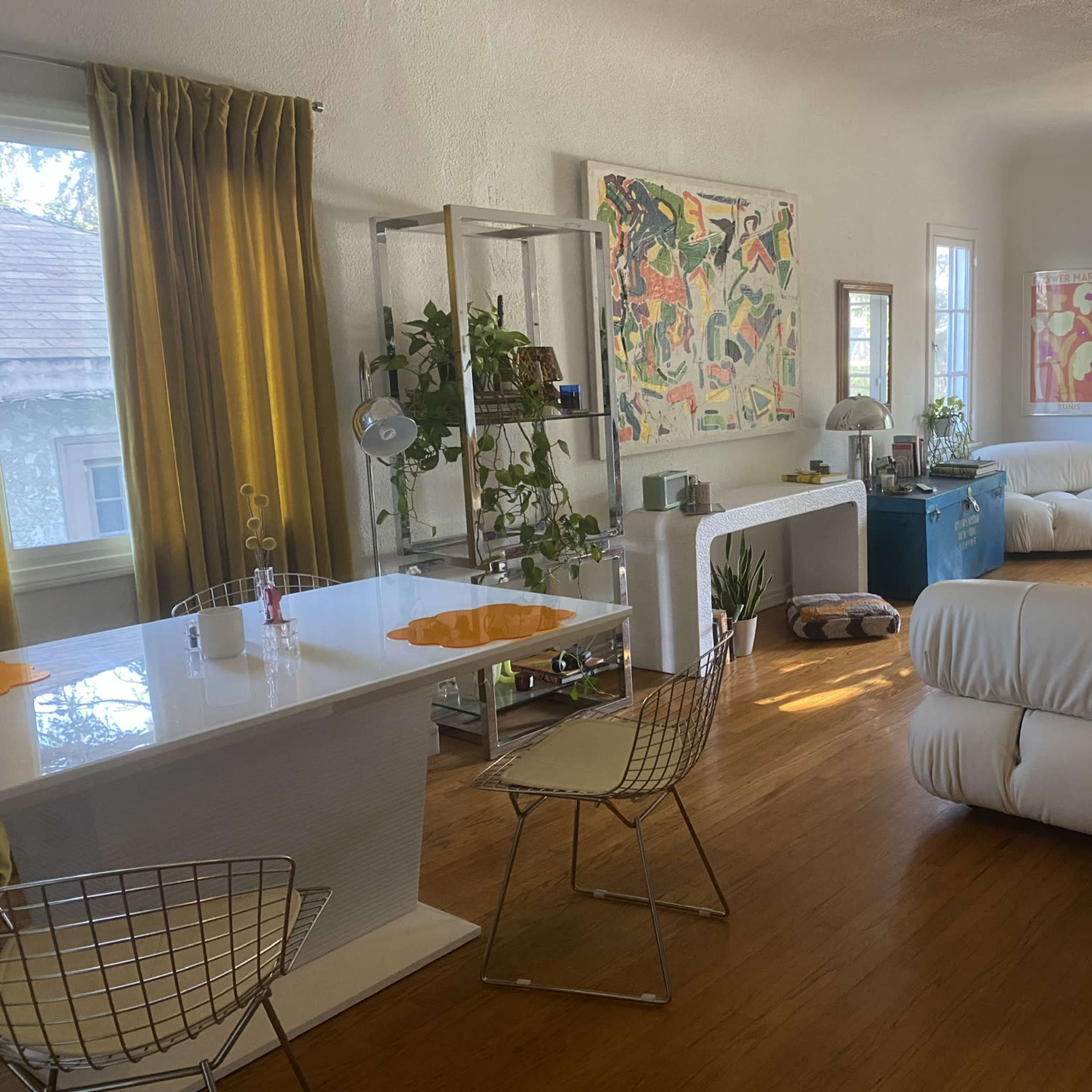 A bright living room with a dining area featuring a white table, metal chairs, and large windows allowing natural light, along with various colorful artworks and furniture pieces.