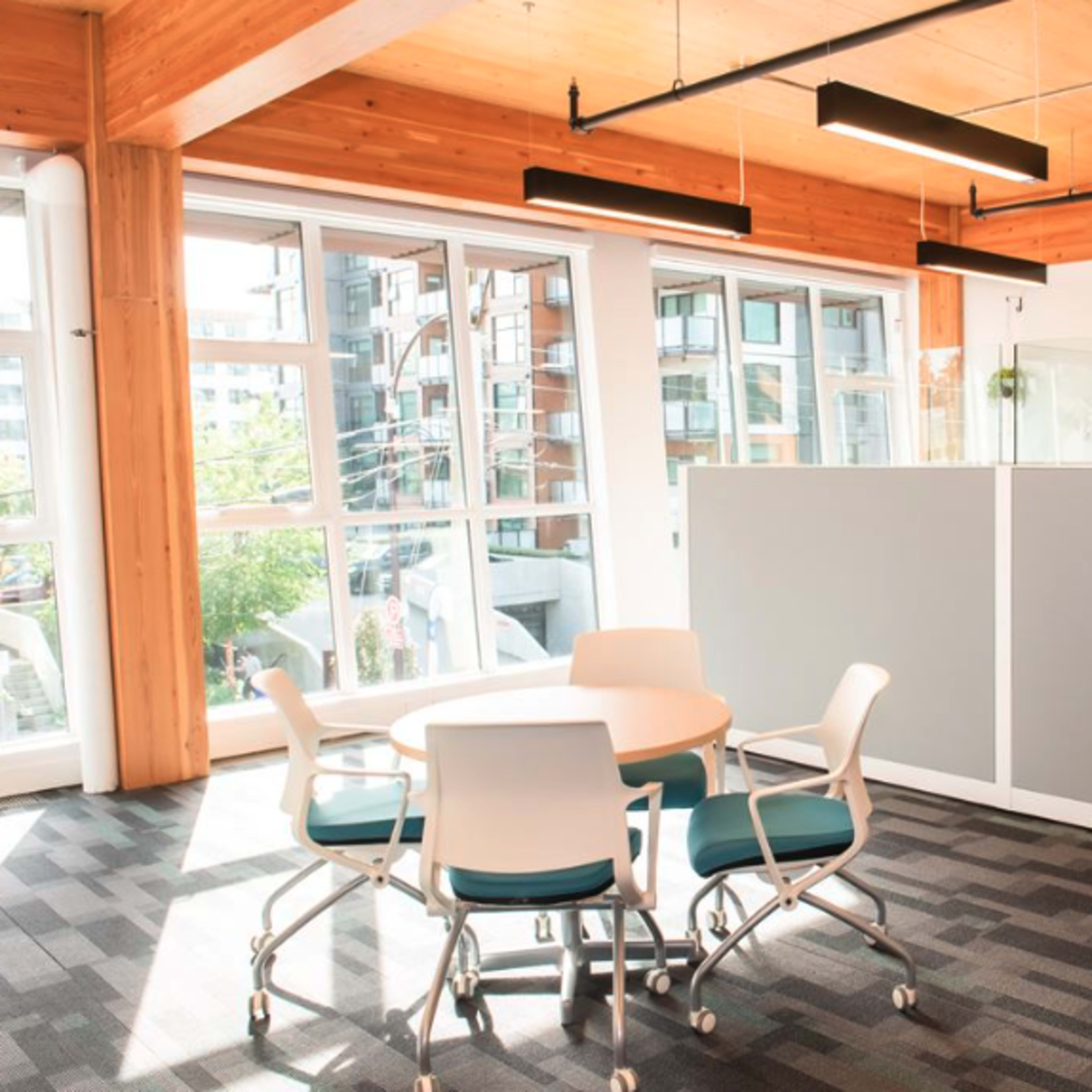 A modern office space features a round table with four chairs next to a large window and a partitioned area behind it.