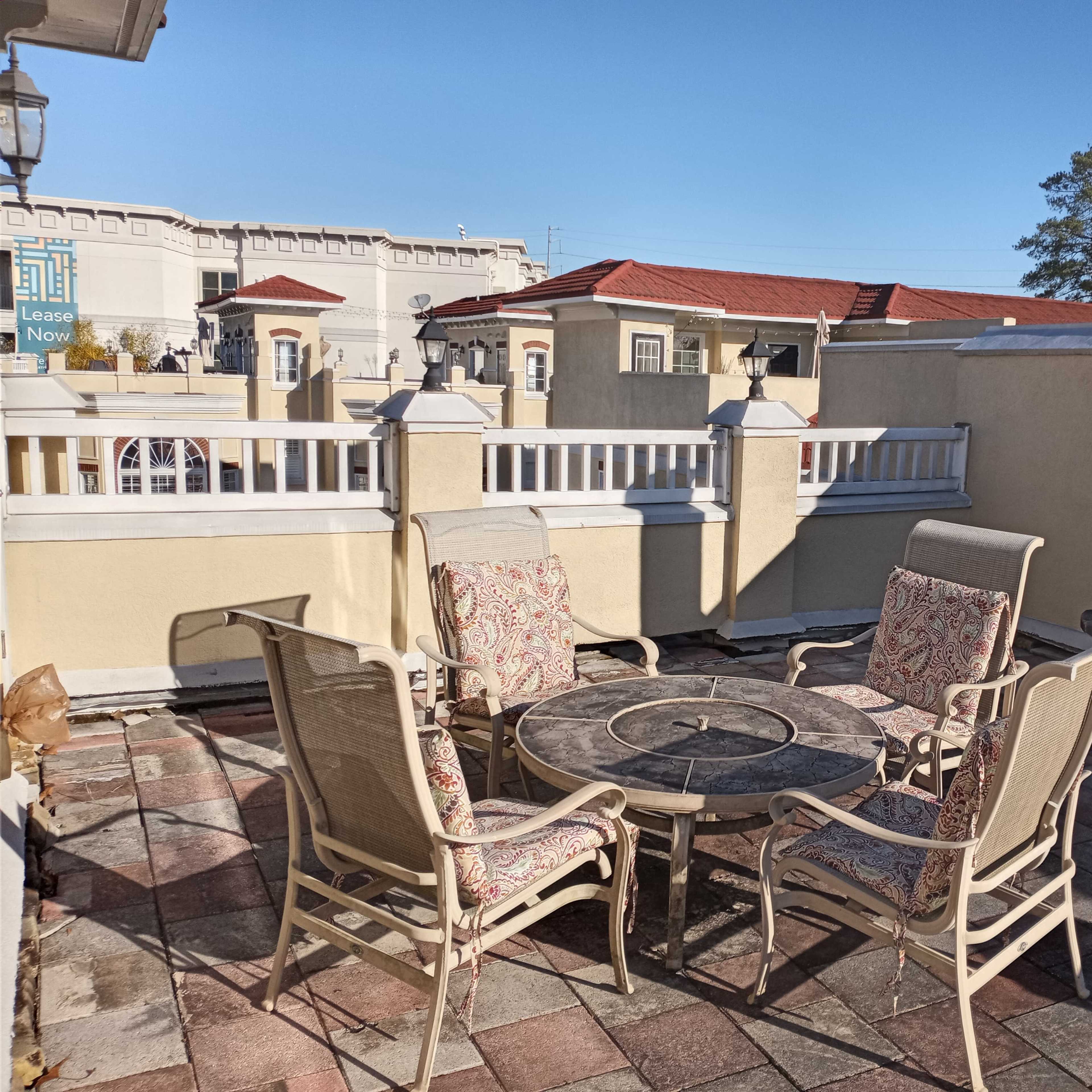 A patio area with four chairs and a round table is set around a stone fireplace, surrounded by a wall with a clear blue sky above.