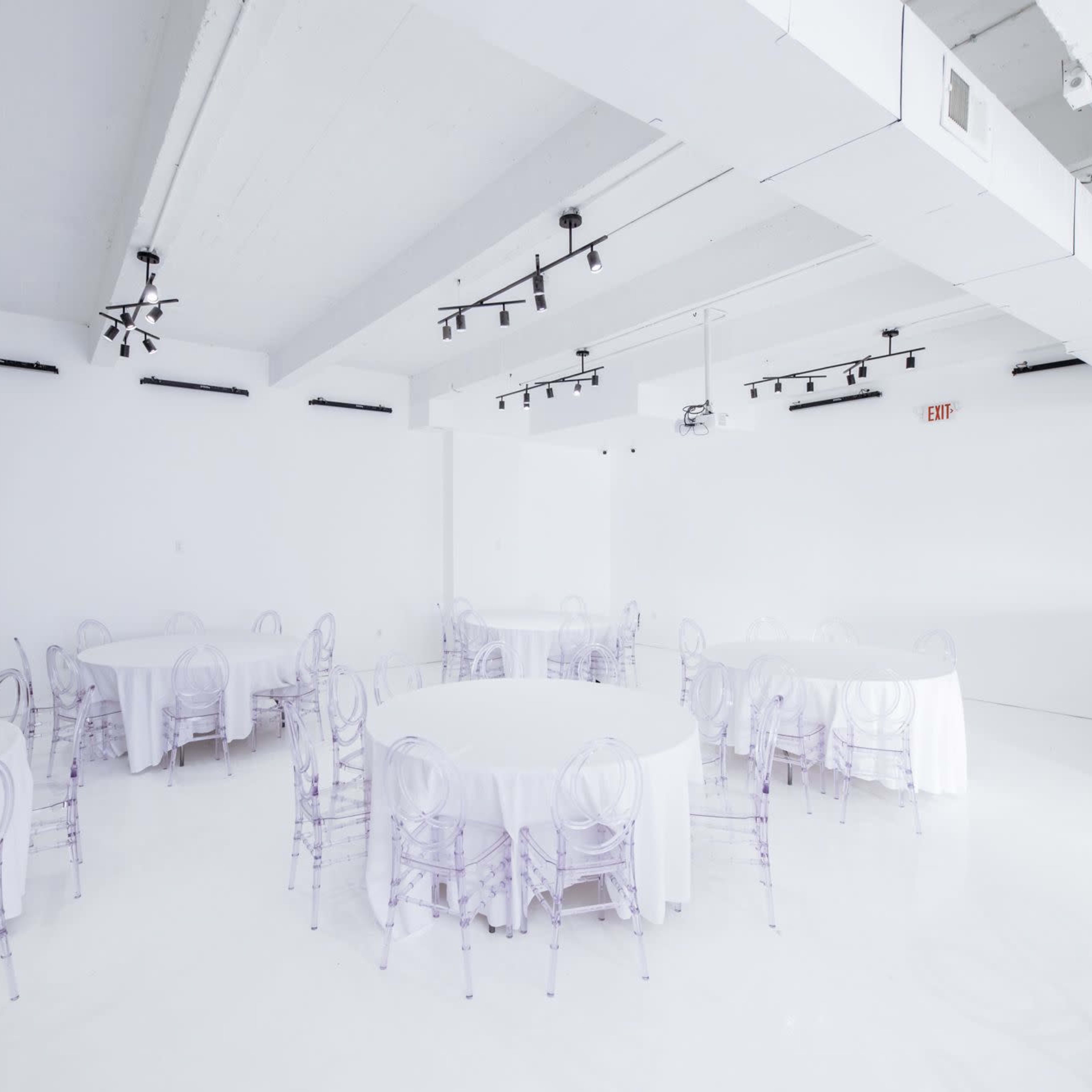 A spacious, minimally decorated white room with several round tables covered in white tablecloths and transparent chairs.