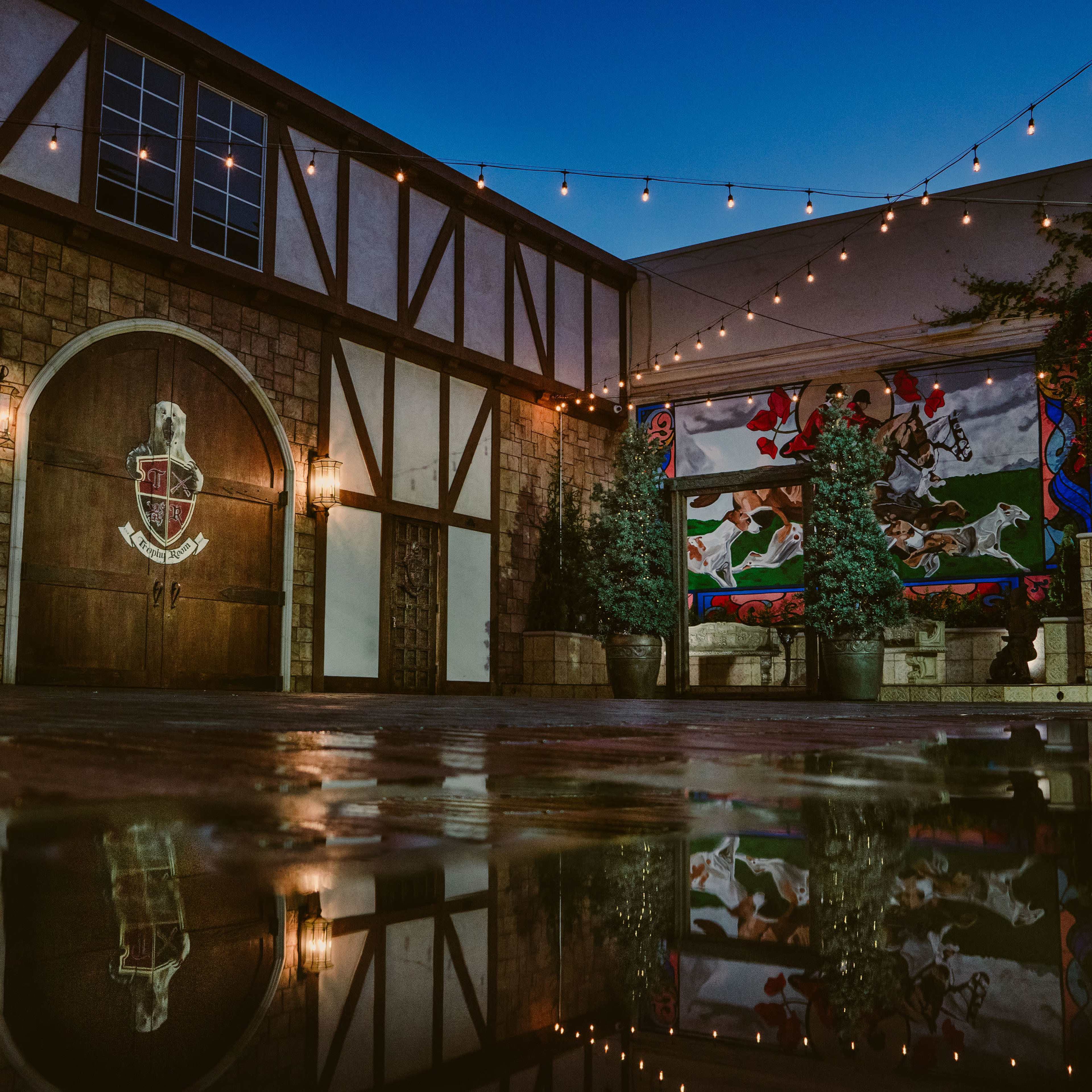 The scene features a rustic courtyard with a stone wall, a large wooden door, and a mural, all illuminated by string lights at dusk, reflecting in a puddle on the ground.