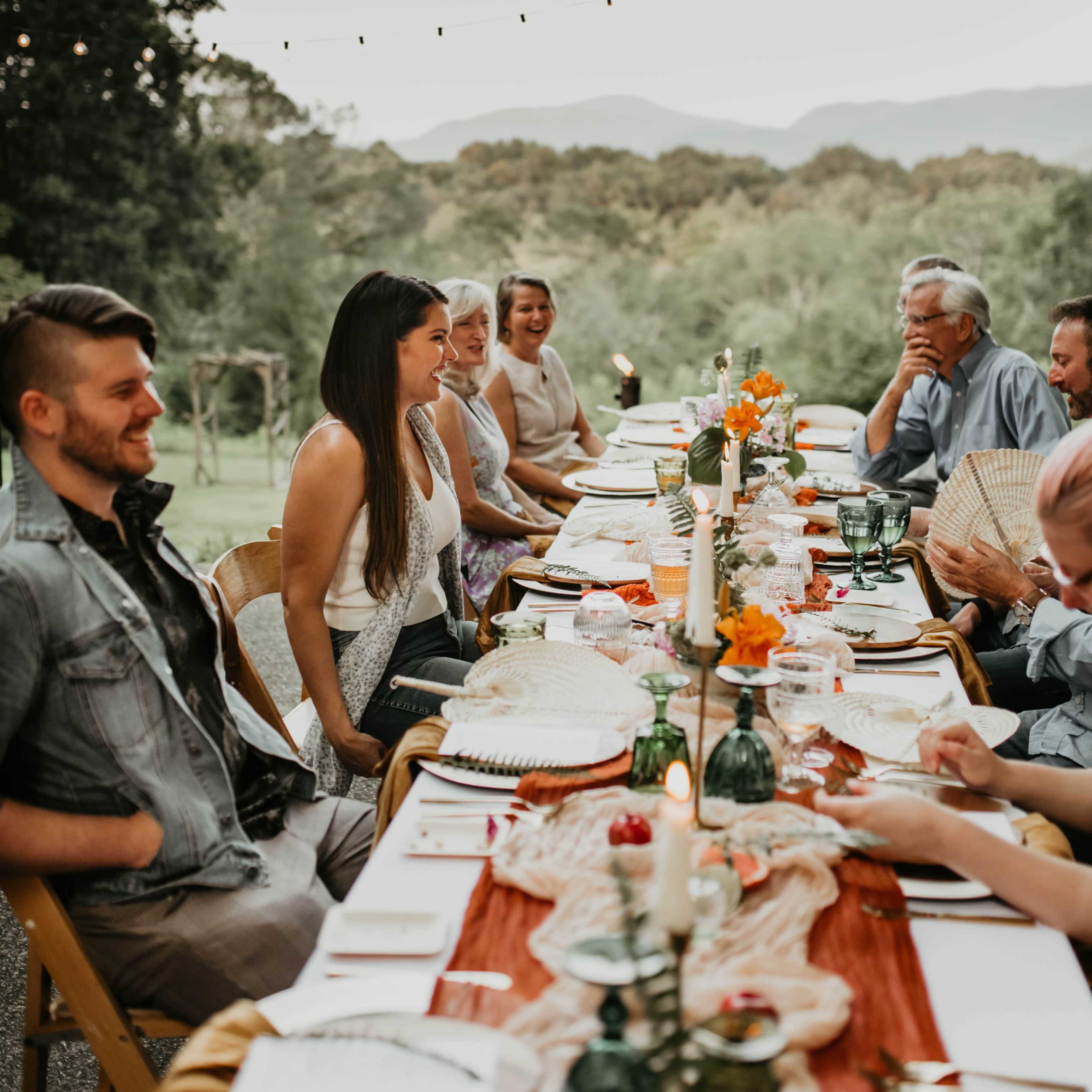 A long table is set for a dinner party outdoors, with multiple guests seated across from each other, surrounded by greenery and mountains in the background.