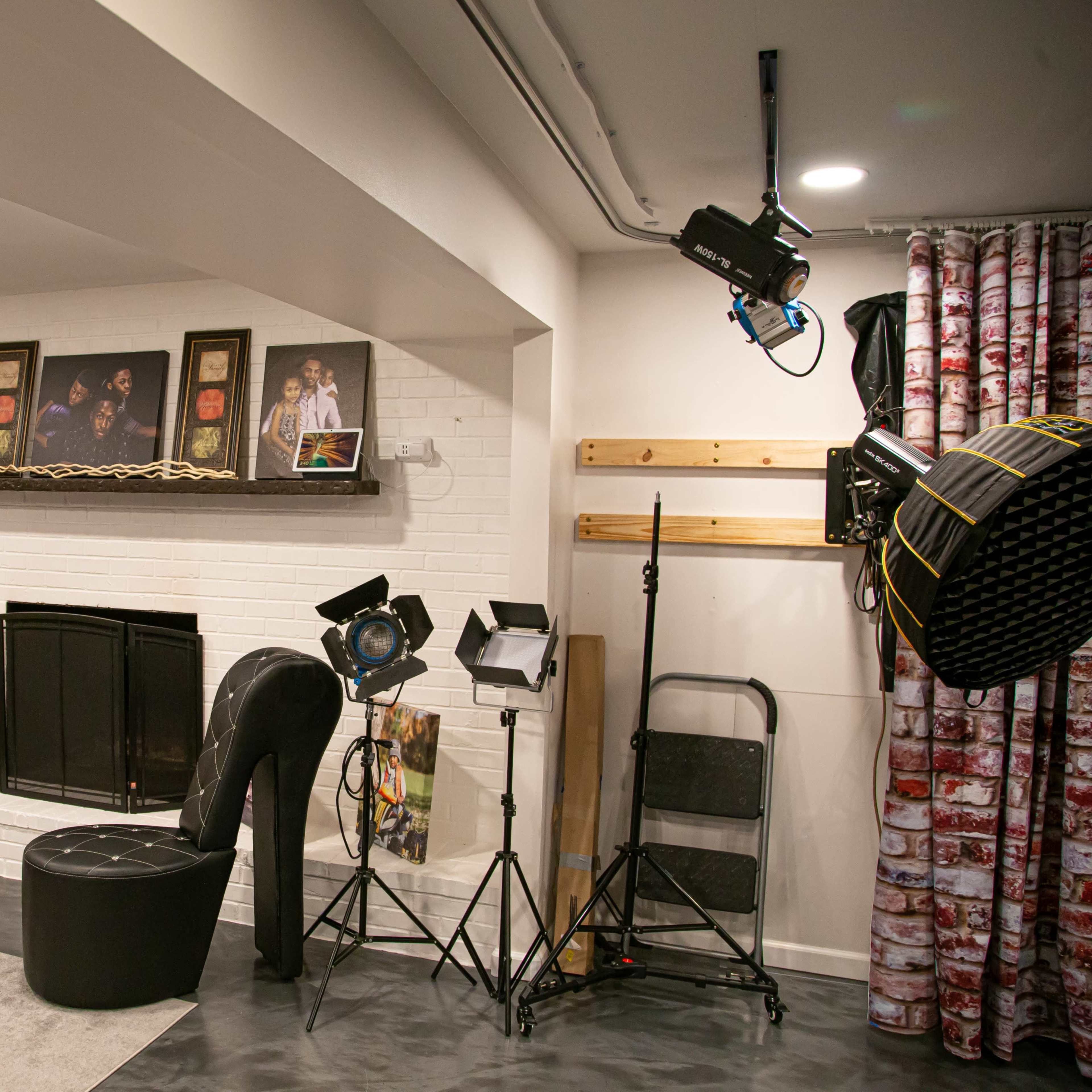 A studio setup featuring a white brick wall with a mantel, various photographs displayed above it, a large high-heeled shoe chair, and studio lighting equipment on the floor.