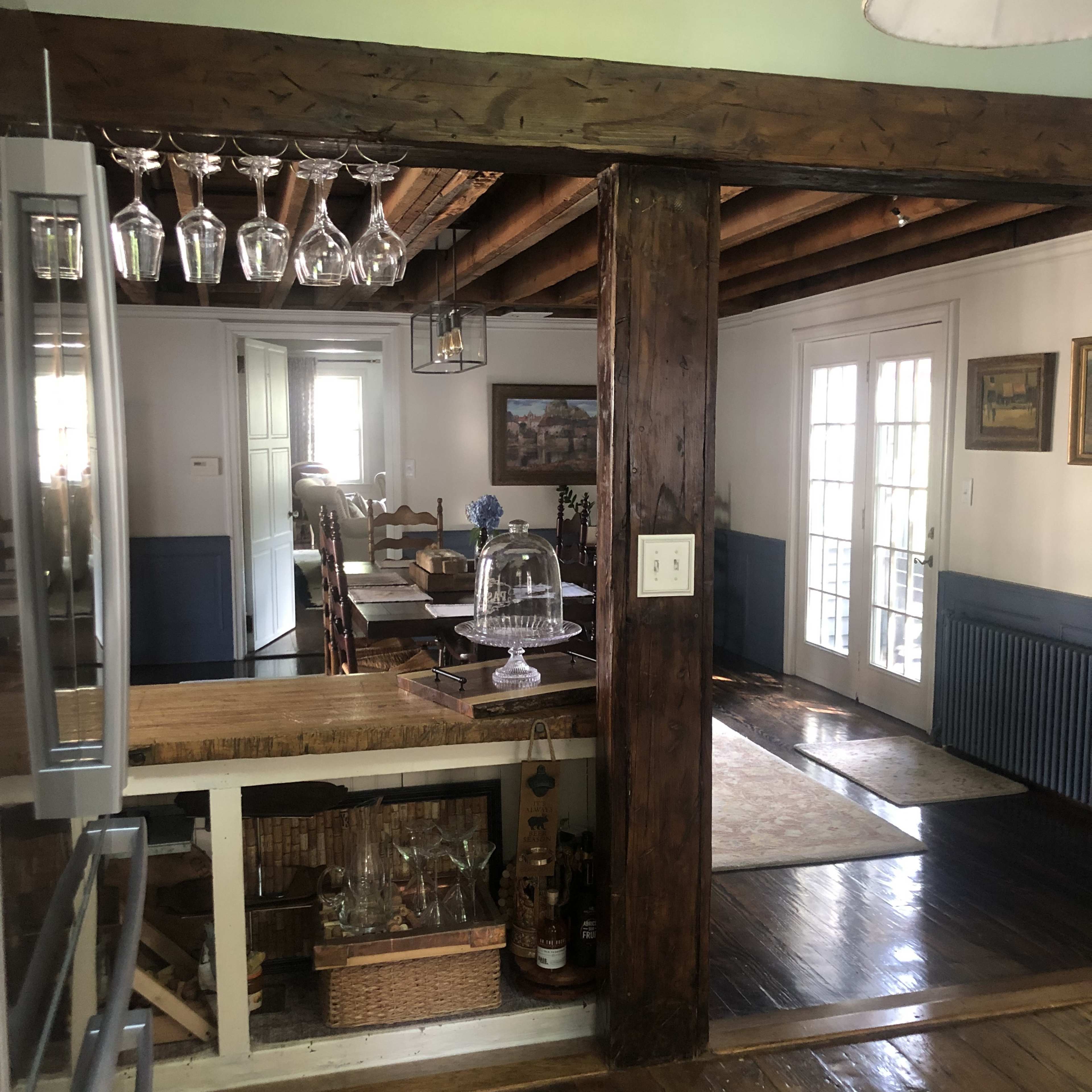 A dining area featuring wooden beams, a glass cabinet, and a dining table with chairs, leading to a brightly lit room with French doors.