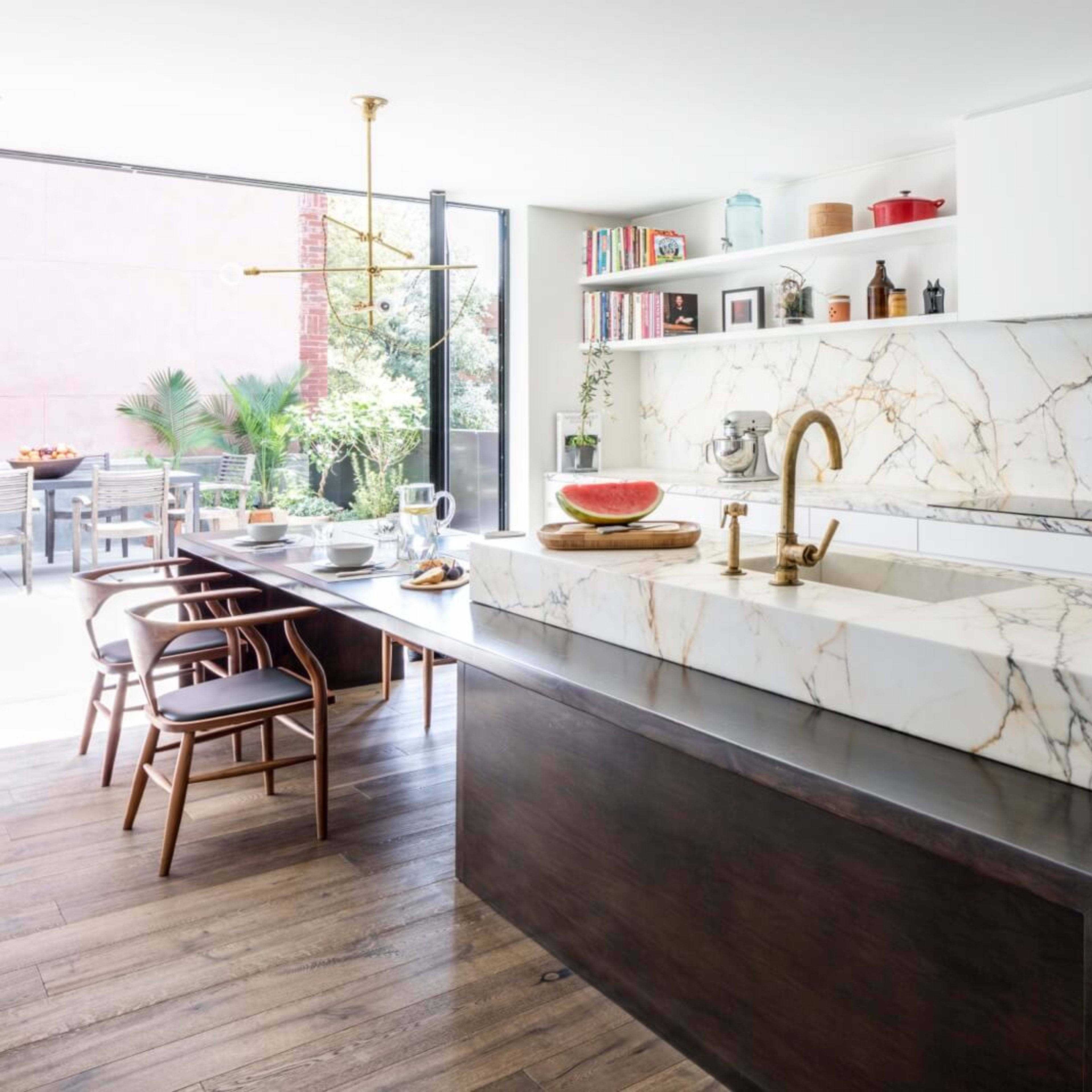 A modern kitchen with a large marble island and a wooden dining table, featuring an outdoor patio visible through large sliding glass doors.