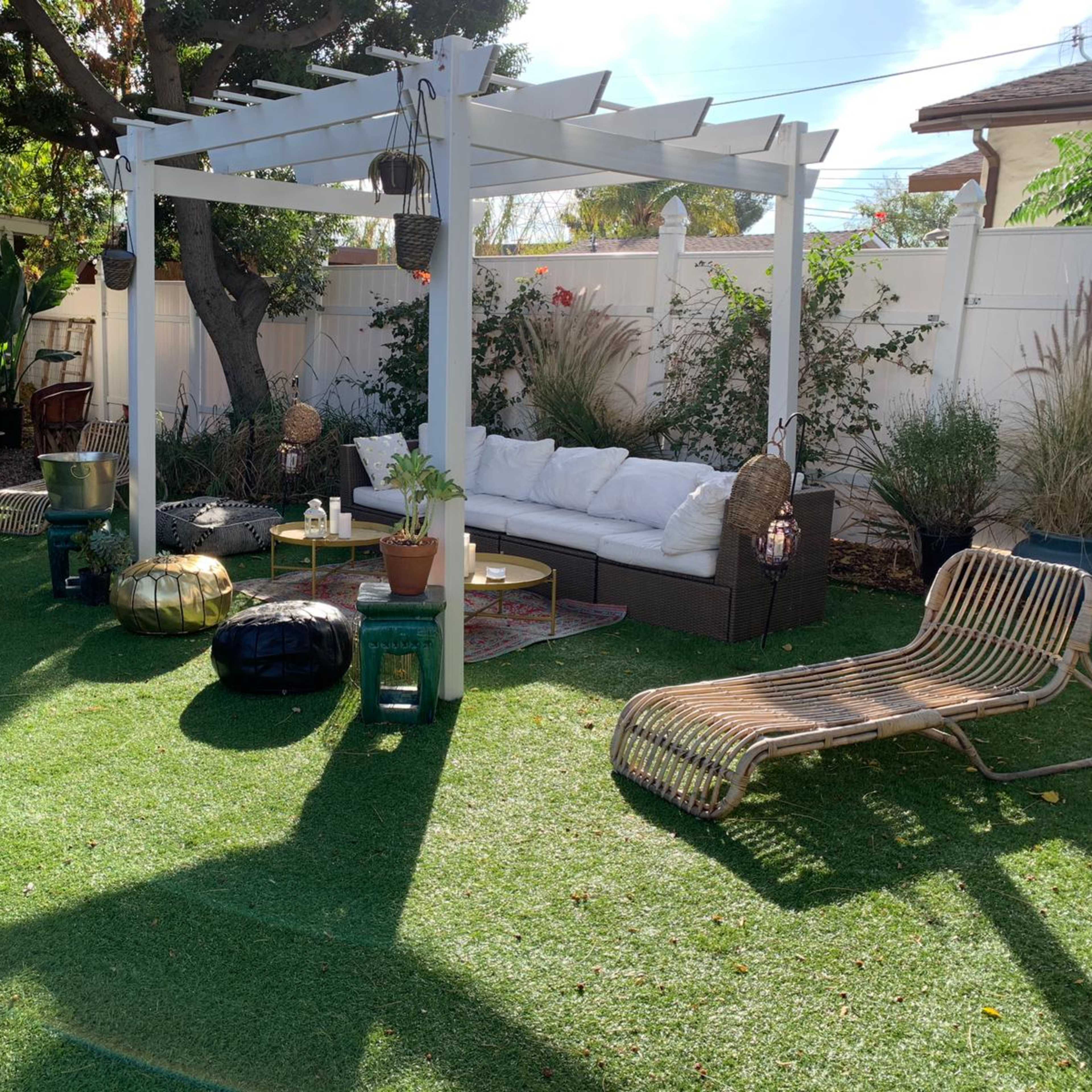 A landscaped backyard with a white pergola, outdoor seating, and various plants on artificial grass.