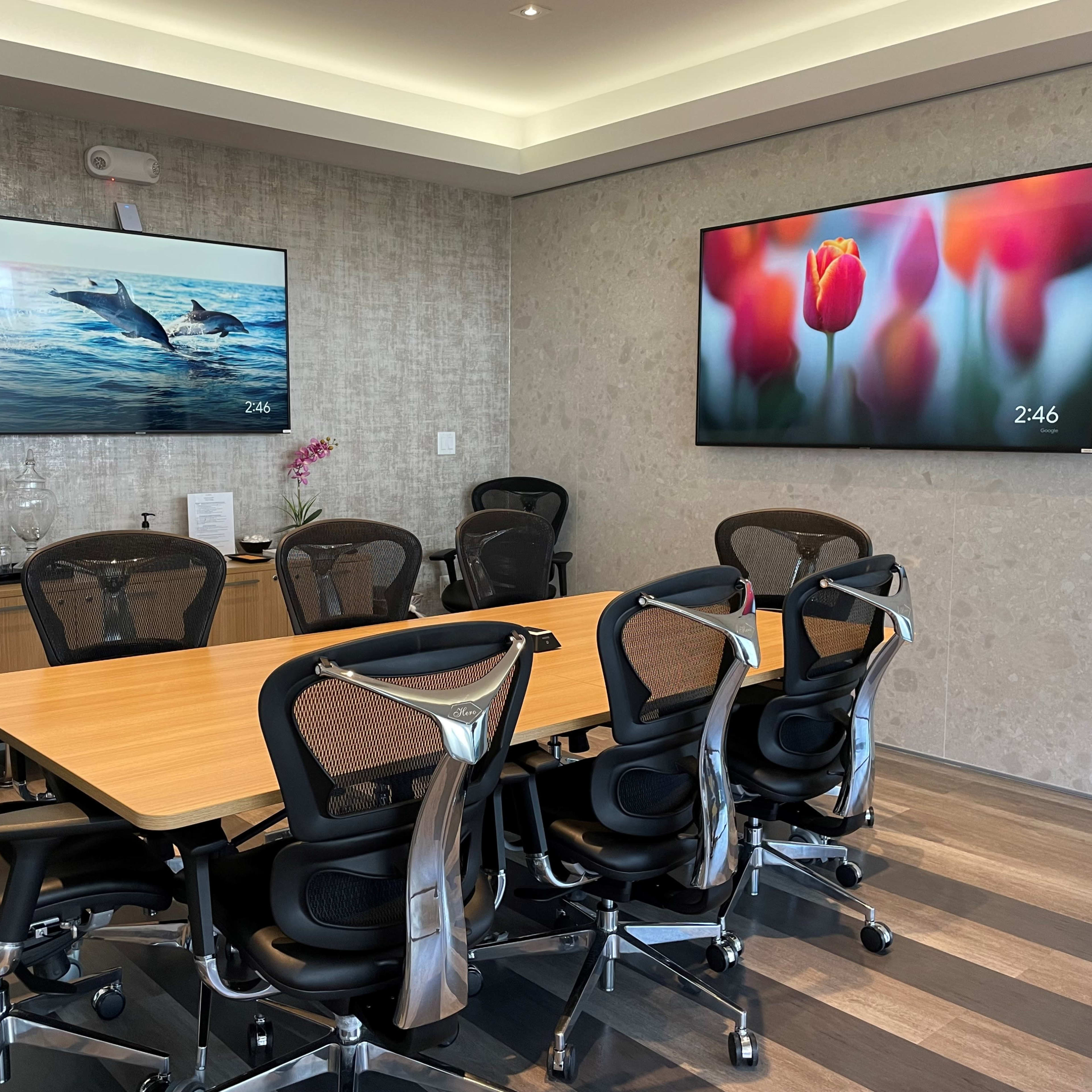 A modern conference room features a large wooden table surrounded by ergonomic chairs, with two screens displaying images on the walls.