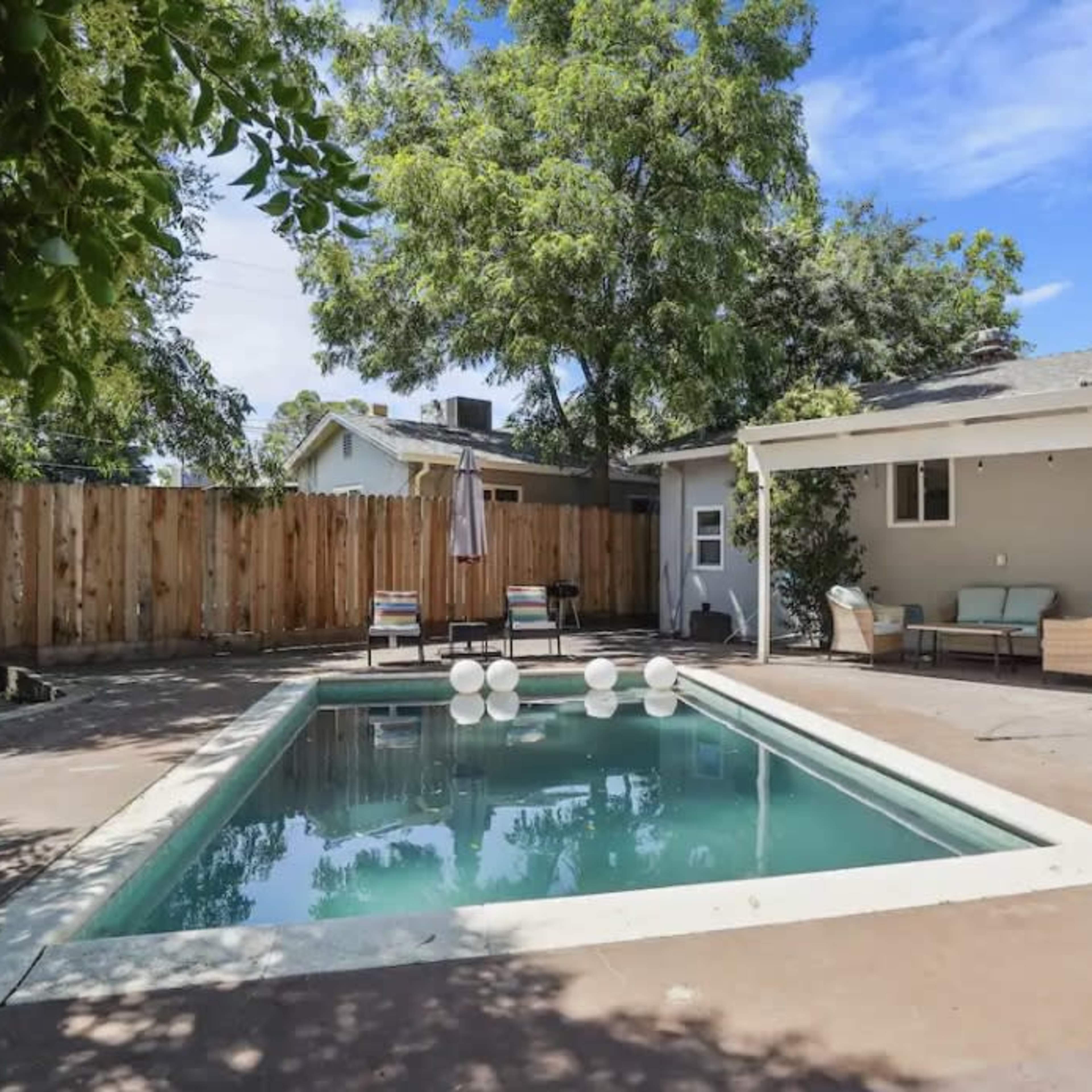 A backyard with a swimming pool surrounded by concrete, patio furniture, and a wooden fence in the background.