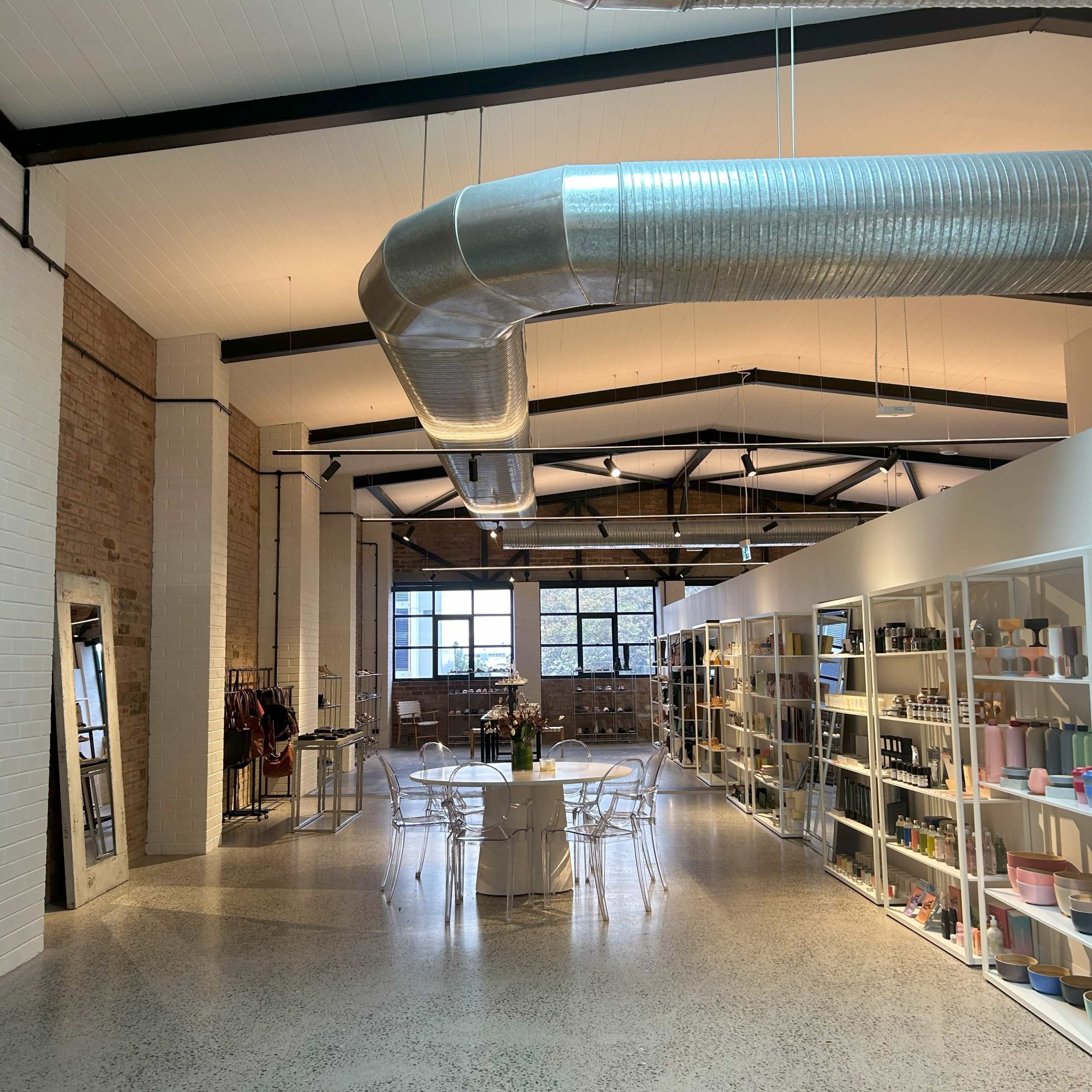 A spacious interior of a contemporary retail store with a large dining table, clear chairs, and shelves filled with various products under industrial-style ductwork.