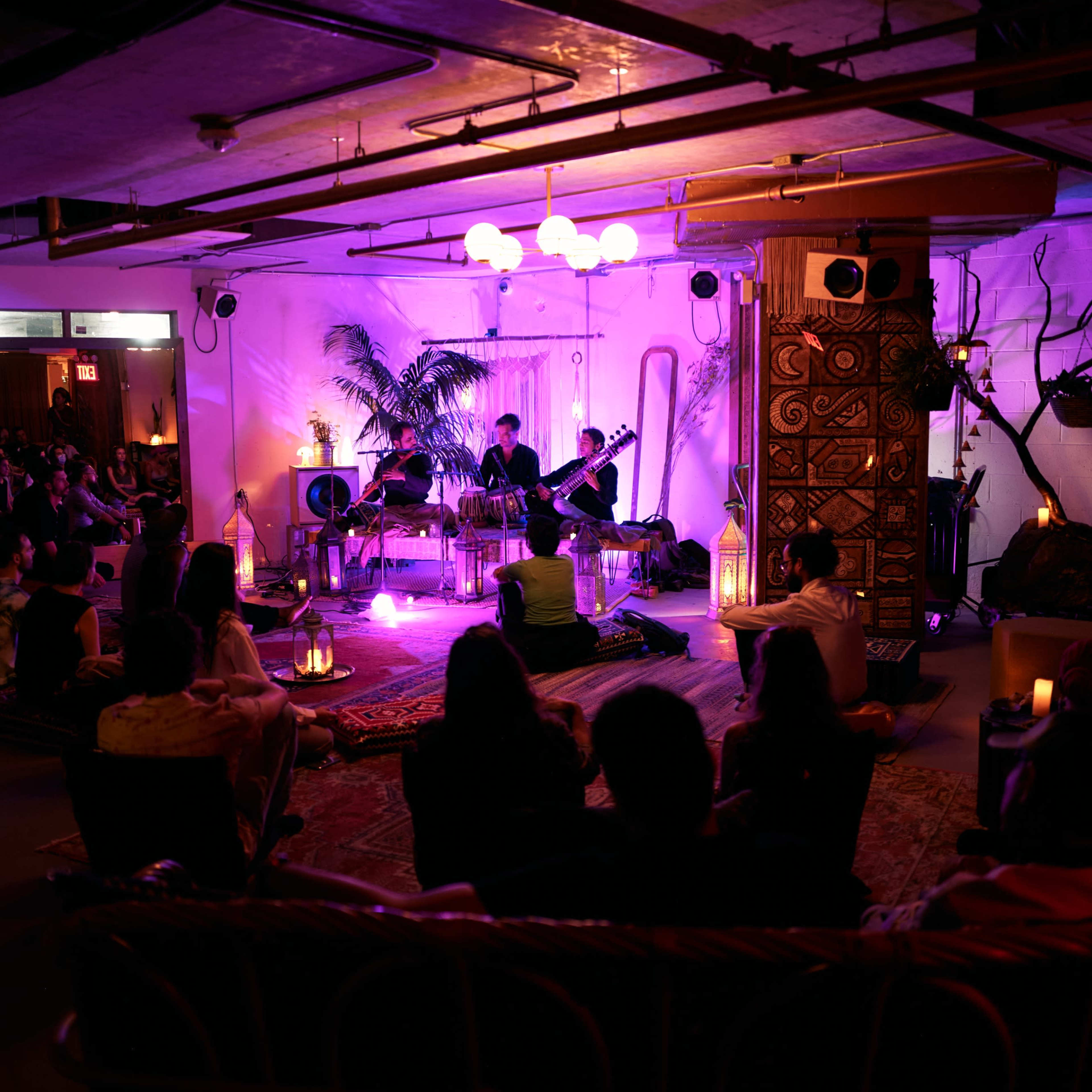 A small audience sits on rugs in a dimly lit room with purple lighting as musicians perform on stage.