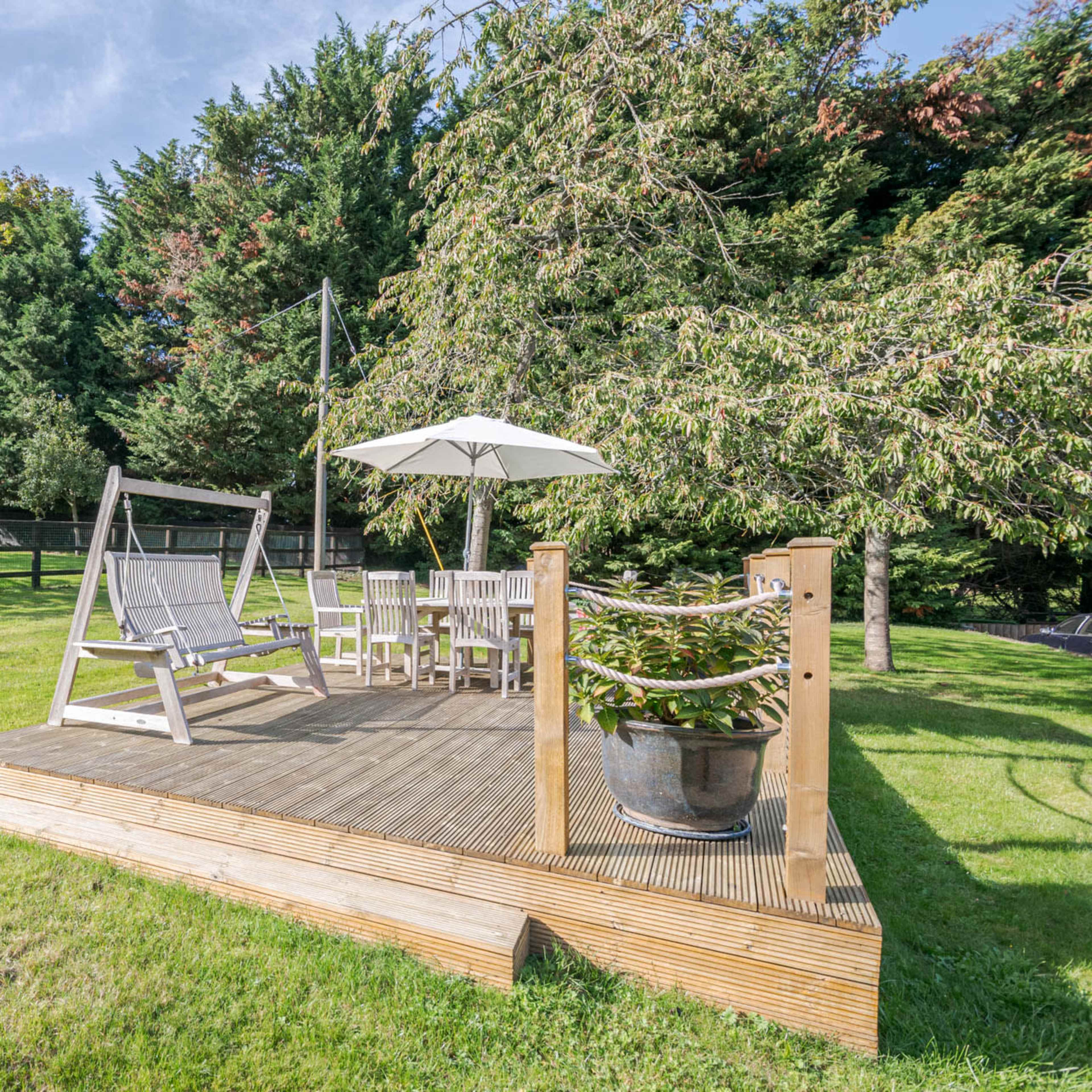A wooden deck with seating, an umbrella, and a swing set in a grassy yard surrounded by trees and a fence.