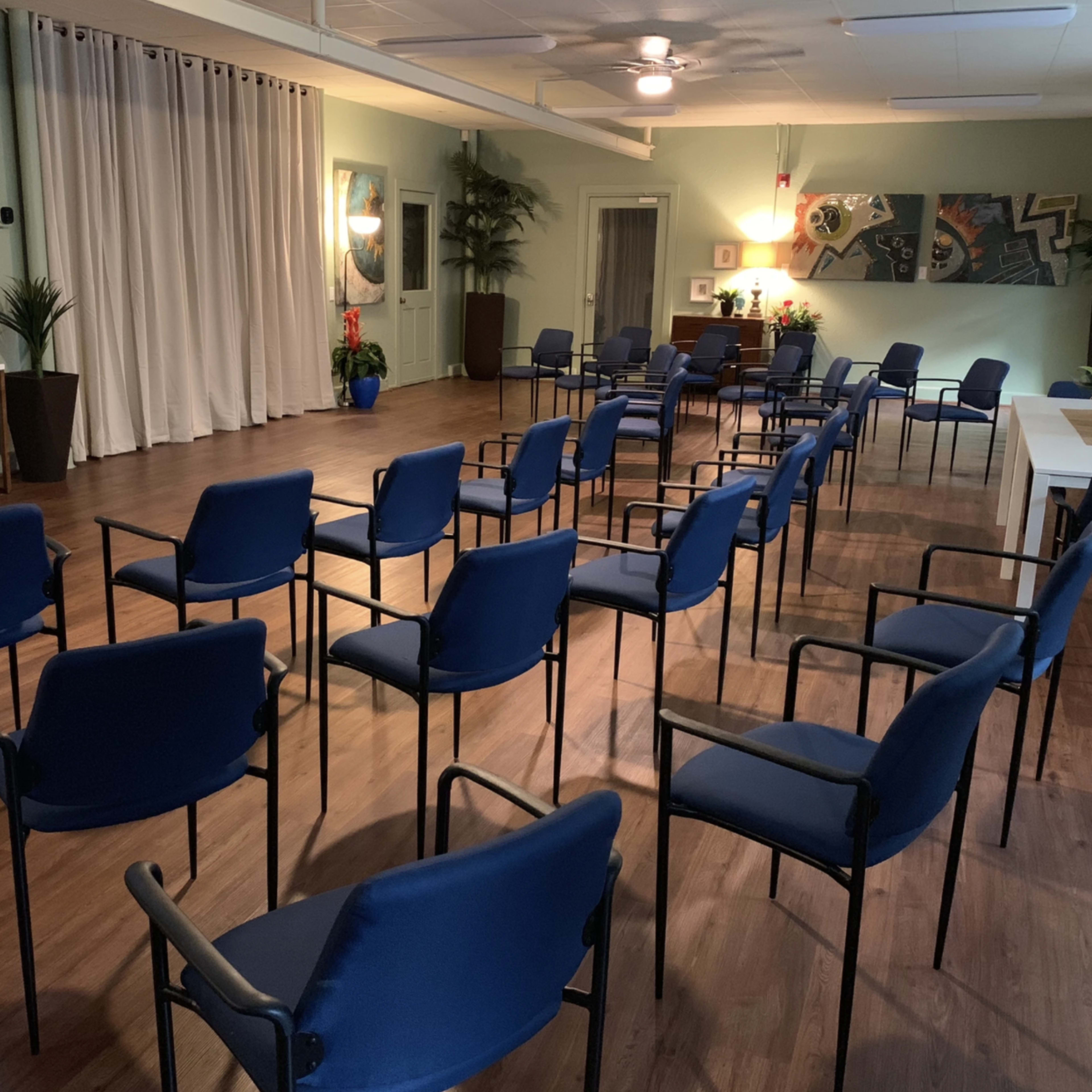 A spacious room with several blue chairs arranged in a circular pattern, surrounded by plants and artwork on the walls.