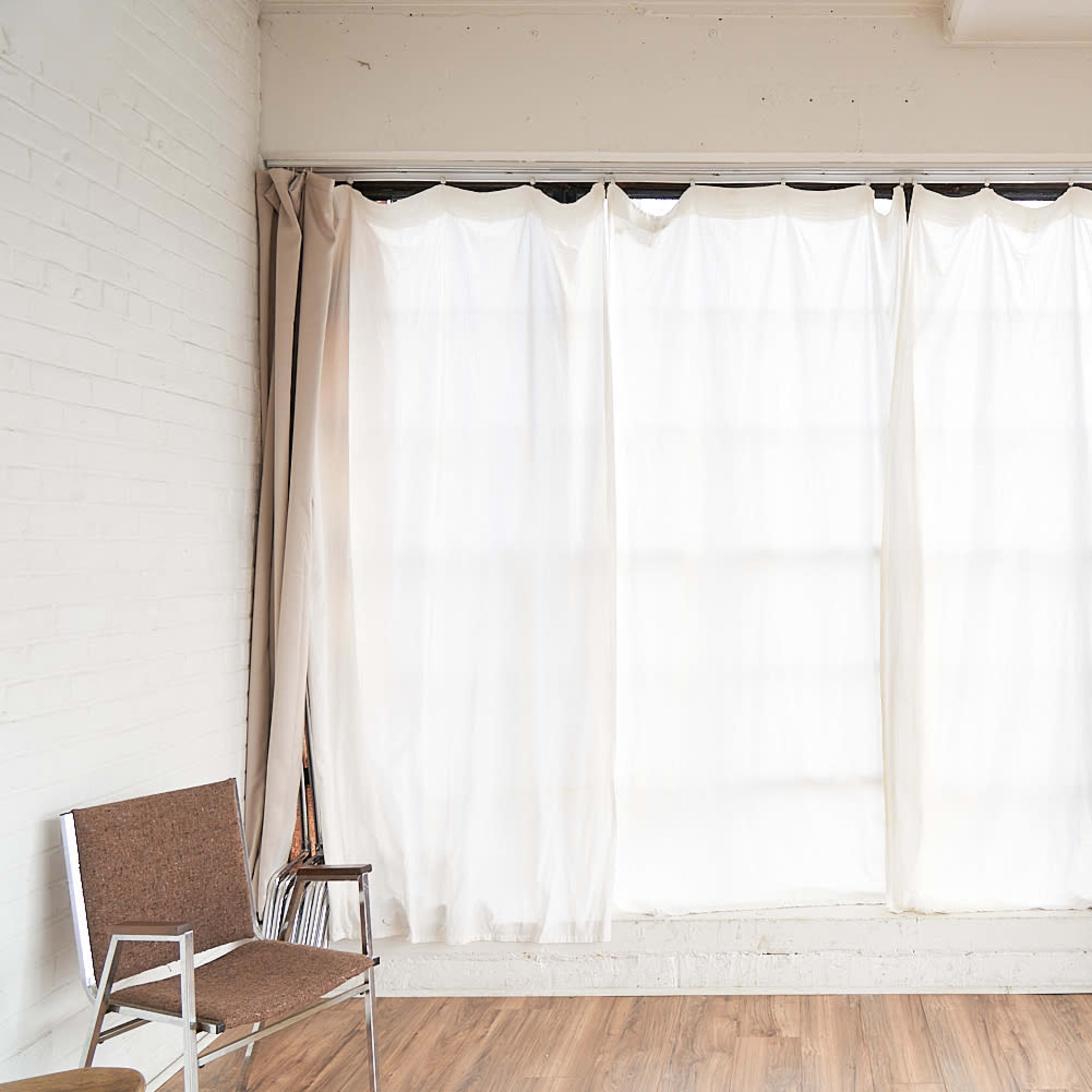 A well-lit room with a large window covered by sheer and solid curtains, alongside a simple brown chair against a white wall.