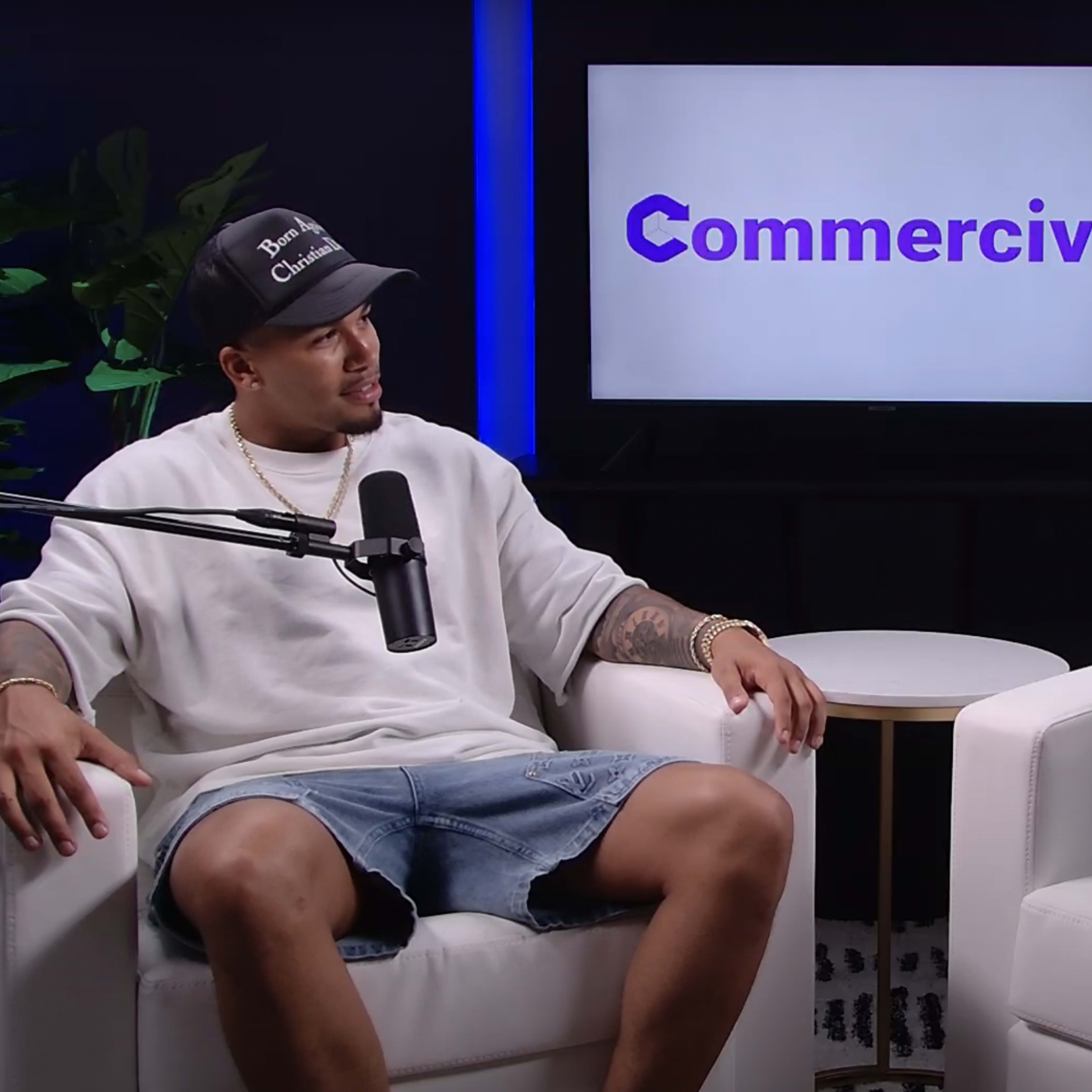 Two individuals are seated in white armchairs in front of a screen displaying the word "Commercial."