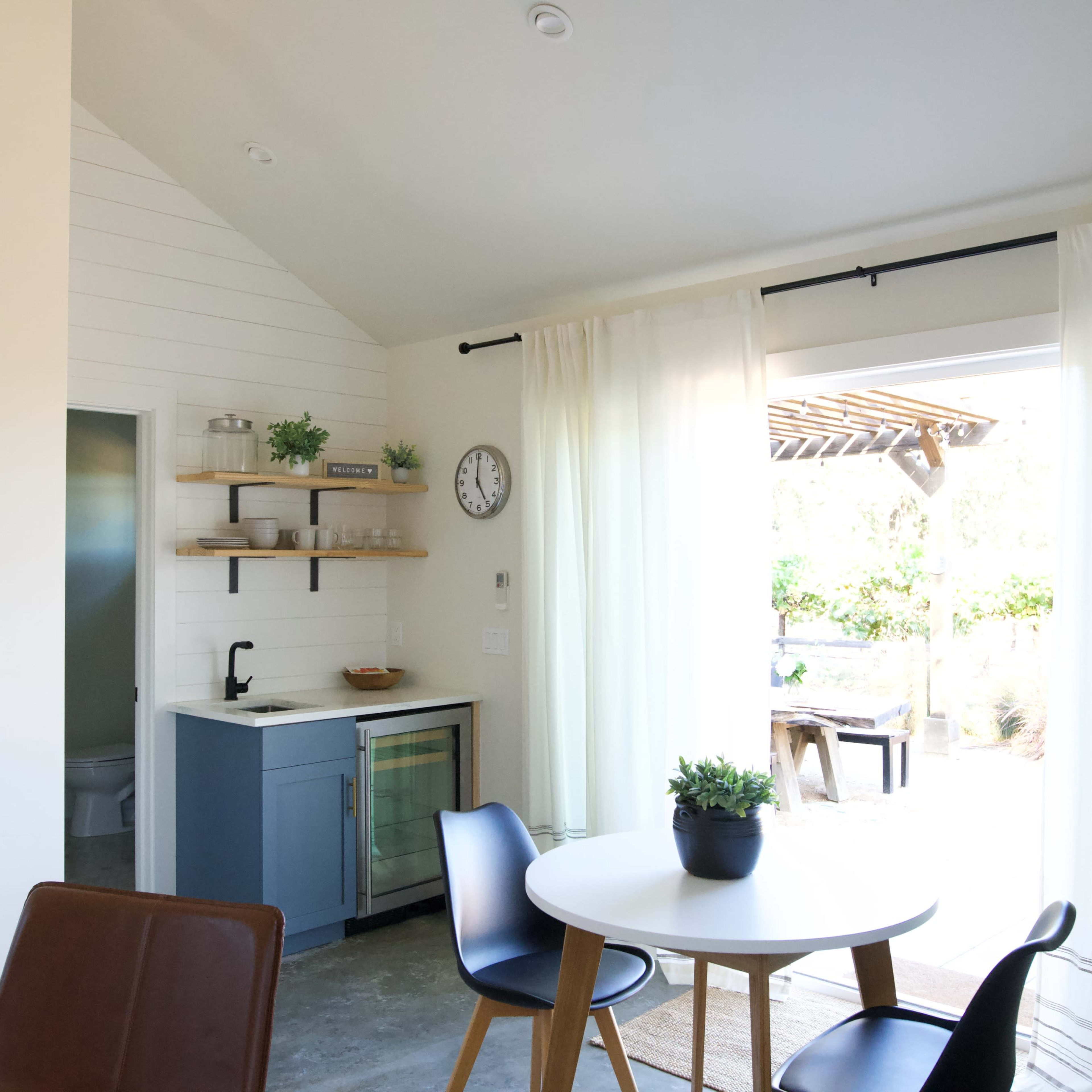 A modern kitchen area features a round table with chairs, open shelving, and large windows that lead to an outdoor space.