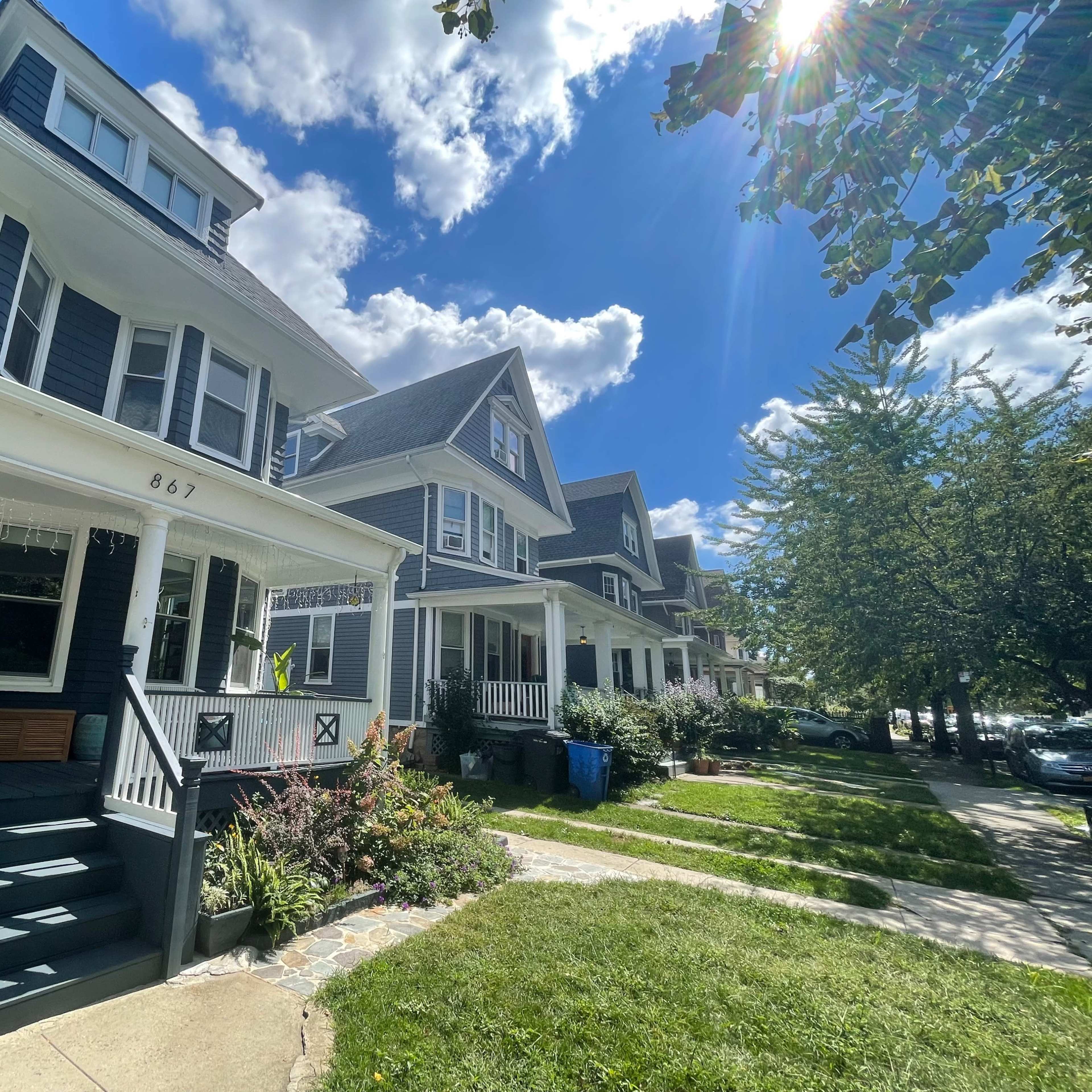 A row of traditional-style houses with porches line a sunny street, accompanied by neatly trimmed lawns and blue trash bins.
