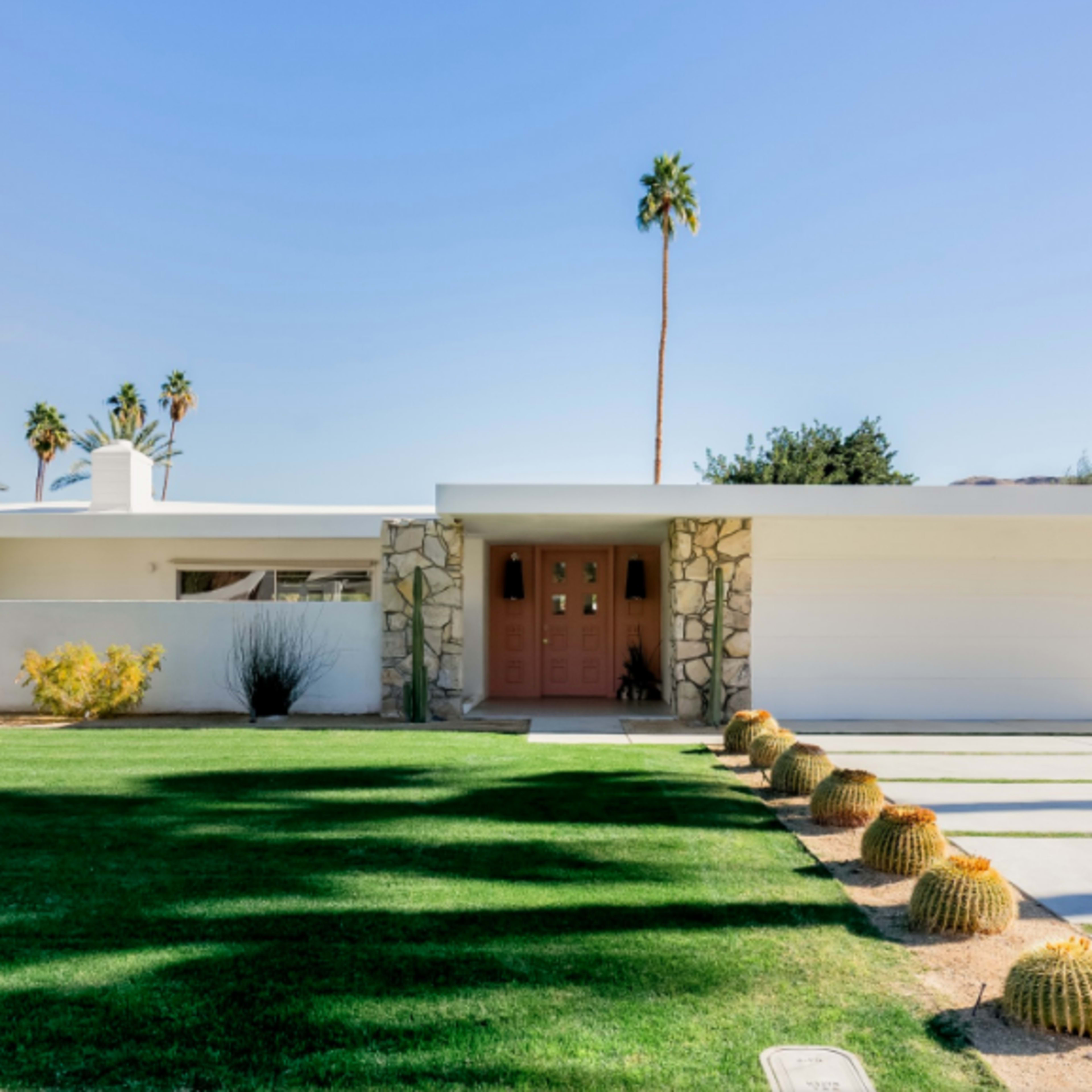 A modern single-story house with a stone-accented entrance, a large garage door, and landscaped front yard featuring cacti and palm trees.