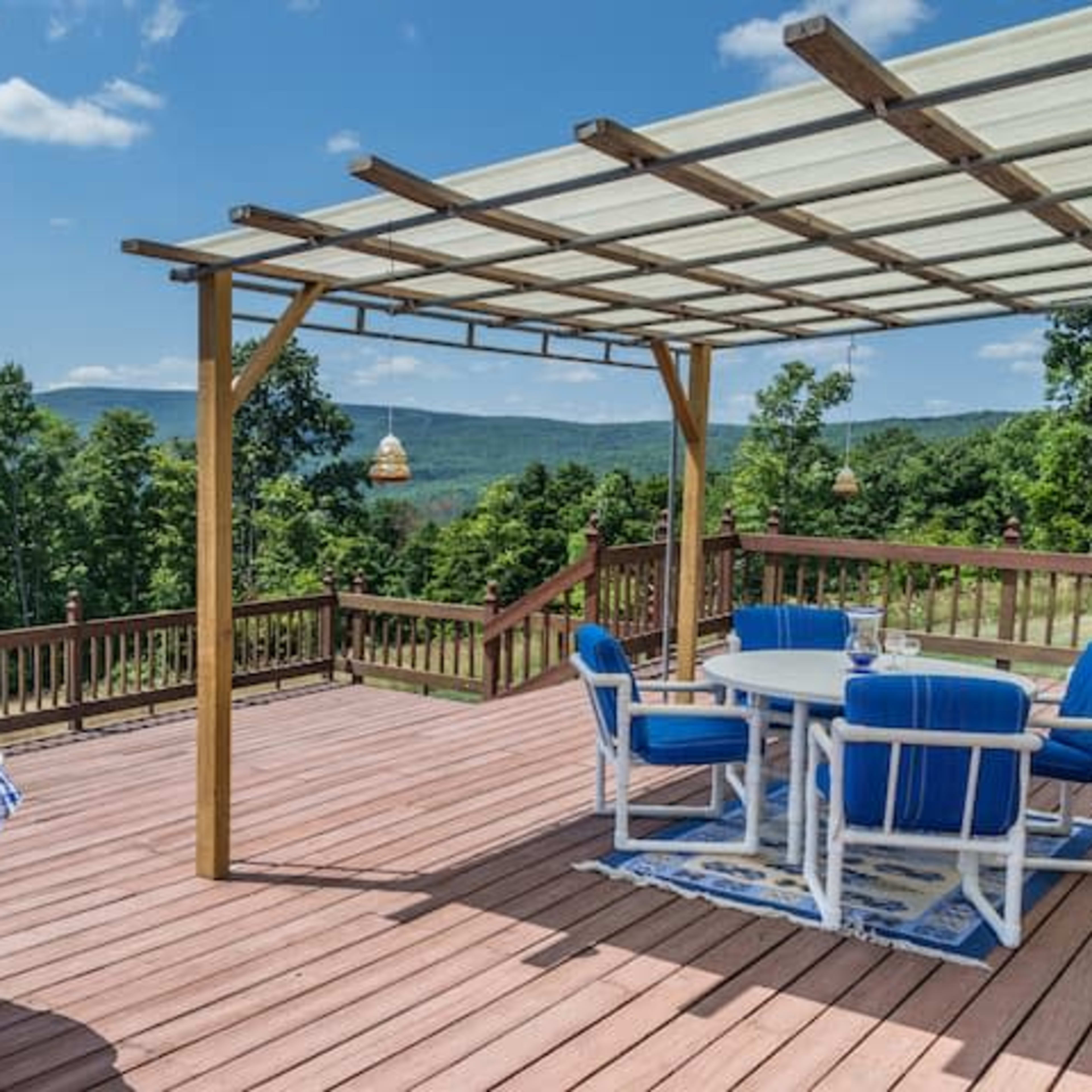 A wooden deck features a dining area with blue and white chairs and tables, surrounded by trees and mountains under a clear blue sky.